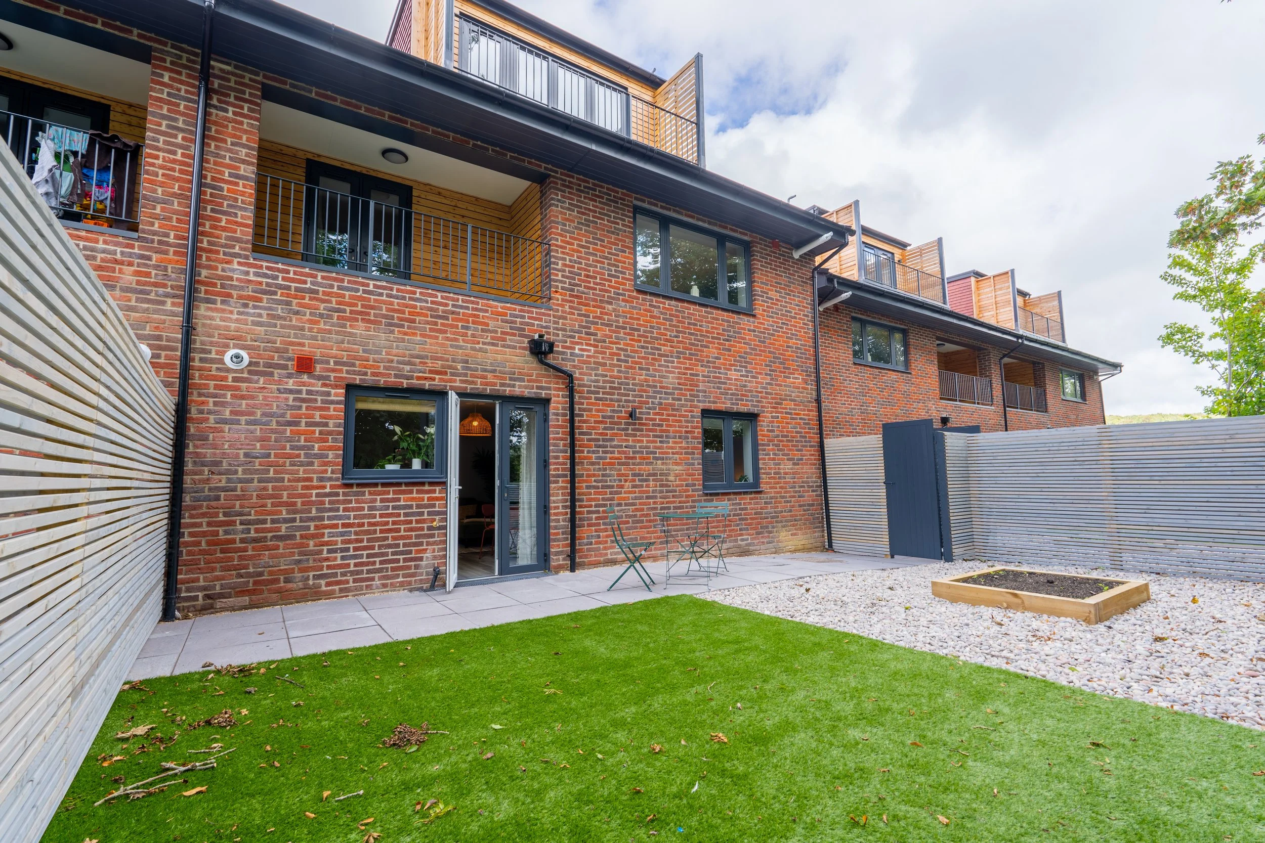 Modern brick building with balconies, sliding glass doors, small patio, artificial grass lawn, and gravel area with planters. Cloudy sky and surrounding greenery.