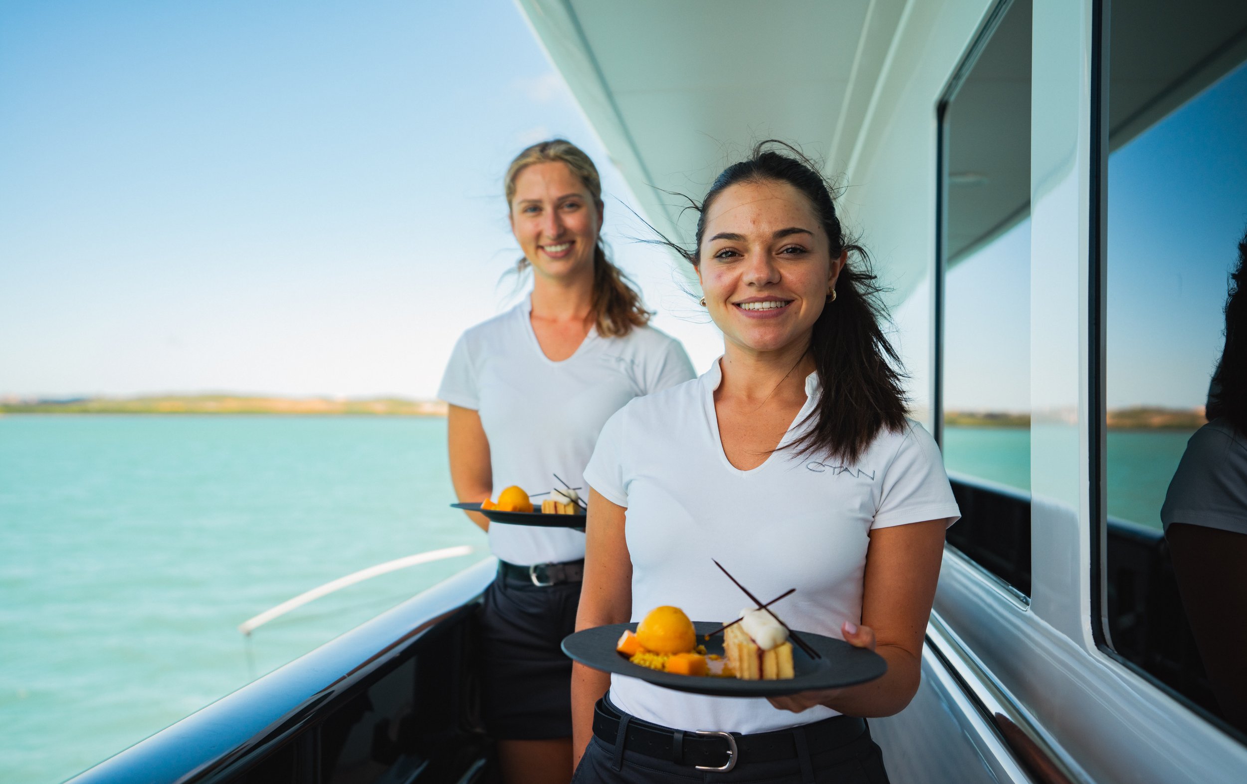 Two women in uniform holding dessert plates on a yacht with ocean and coastline in the background.
