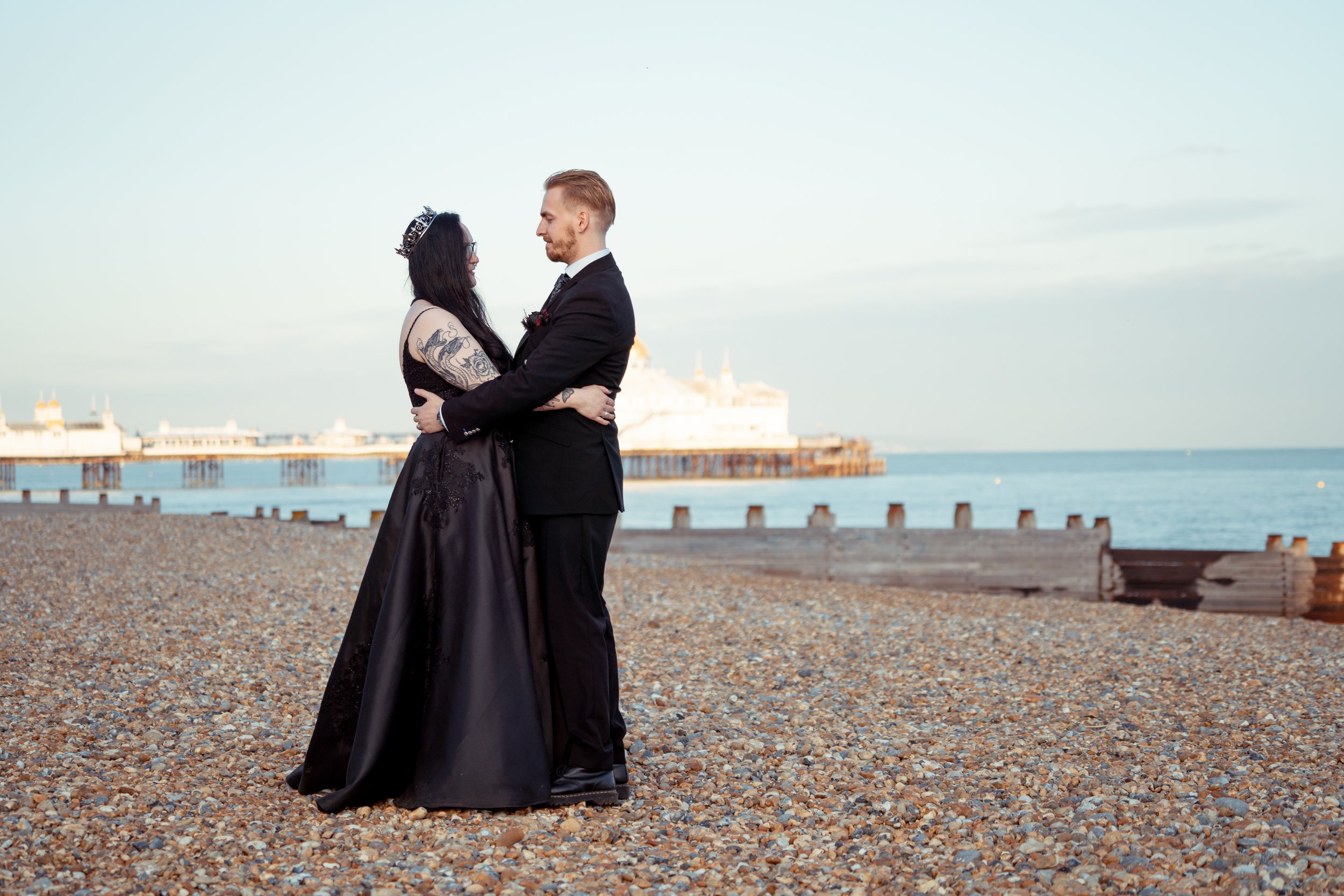 A couple in formal attire embracing on a pebble beach with a pier in the background.