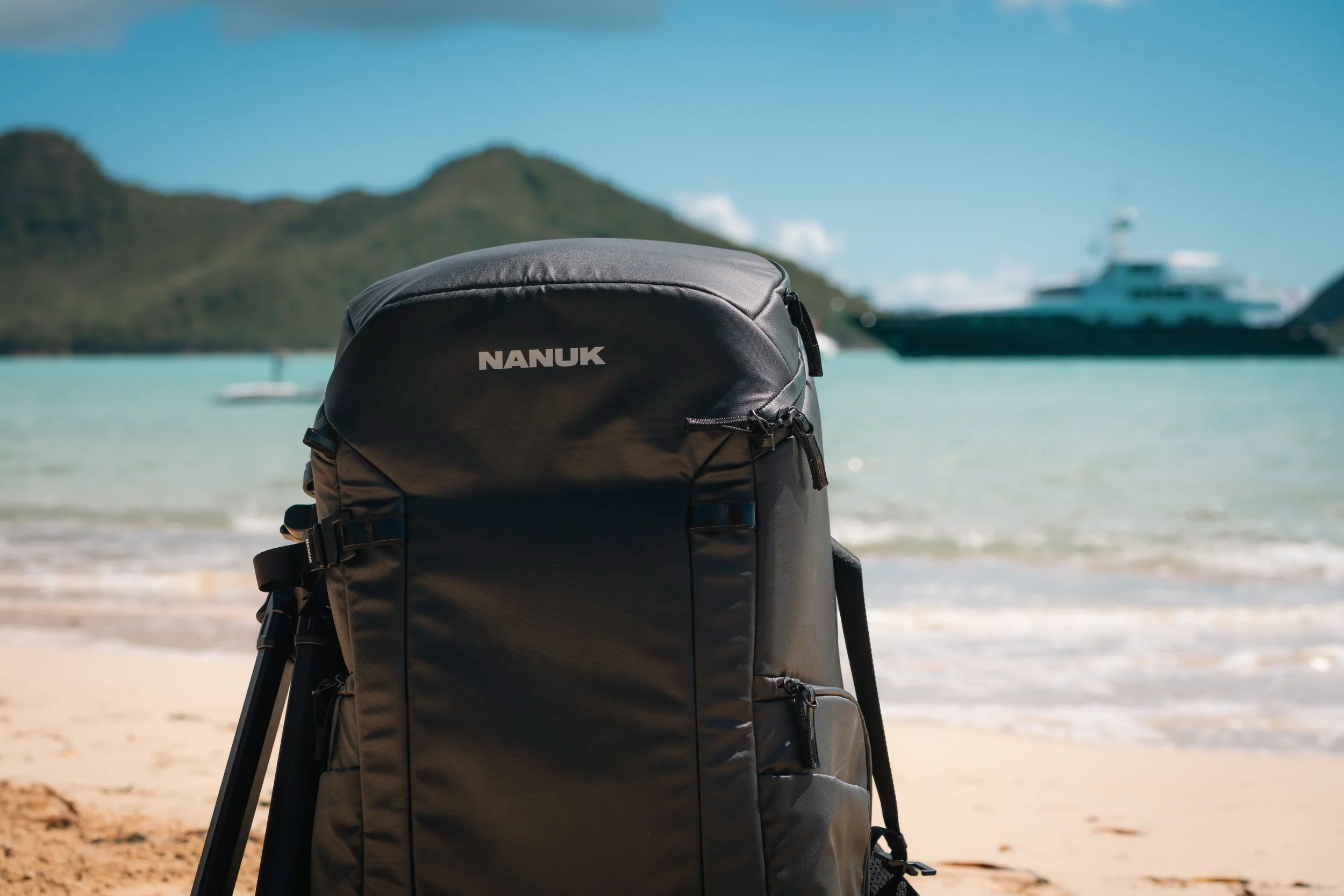 Black Nanuk backpack on a sandy beach with mountains and a ship in the background.