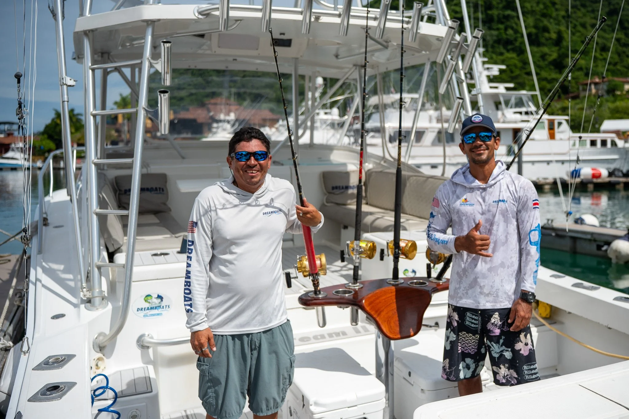 Two men stand on a boat docked at a marina, holding fishing rods and smiling at the camera, with other boats and green hills in the background.