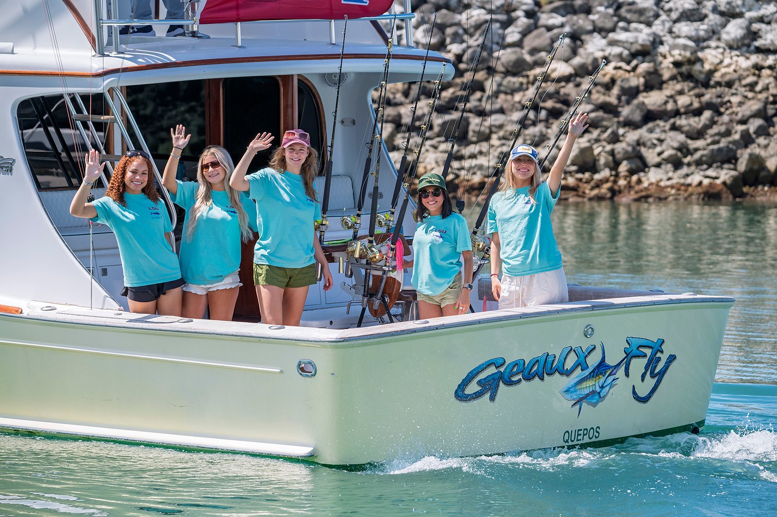 Five women on a boat called 'Geryx Fly' near a rocky shoreline, waving and smiling during daytime. They are dressed in casual summer clothing, some wearing hats and sunglasses, with fishing rods mounted on the boat.