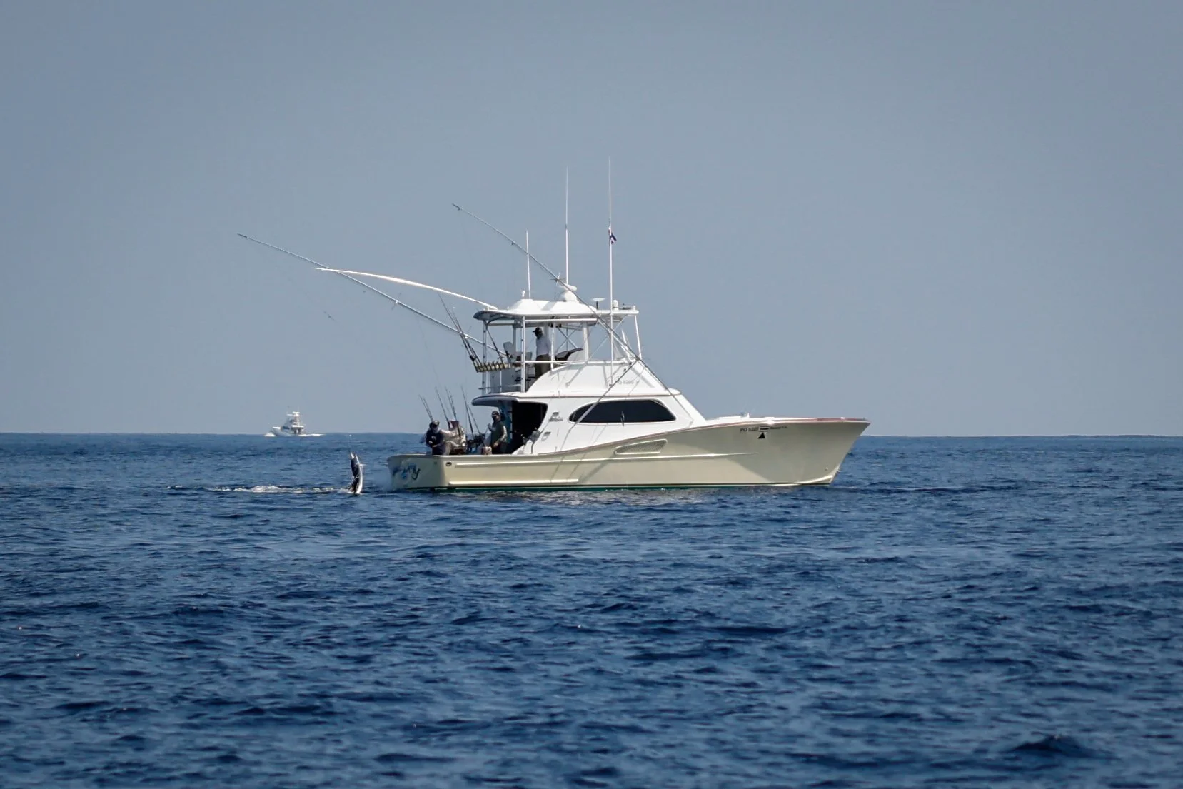 A white yacht on calm ocean waters with two people visible on deck and fishing rods attached to the sides. Two other boats are visible in the background under a clear sky.