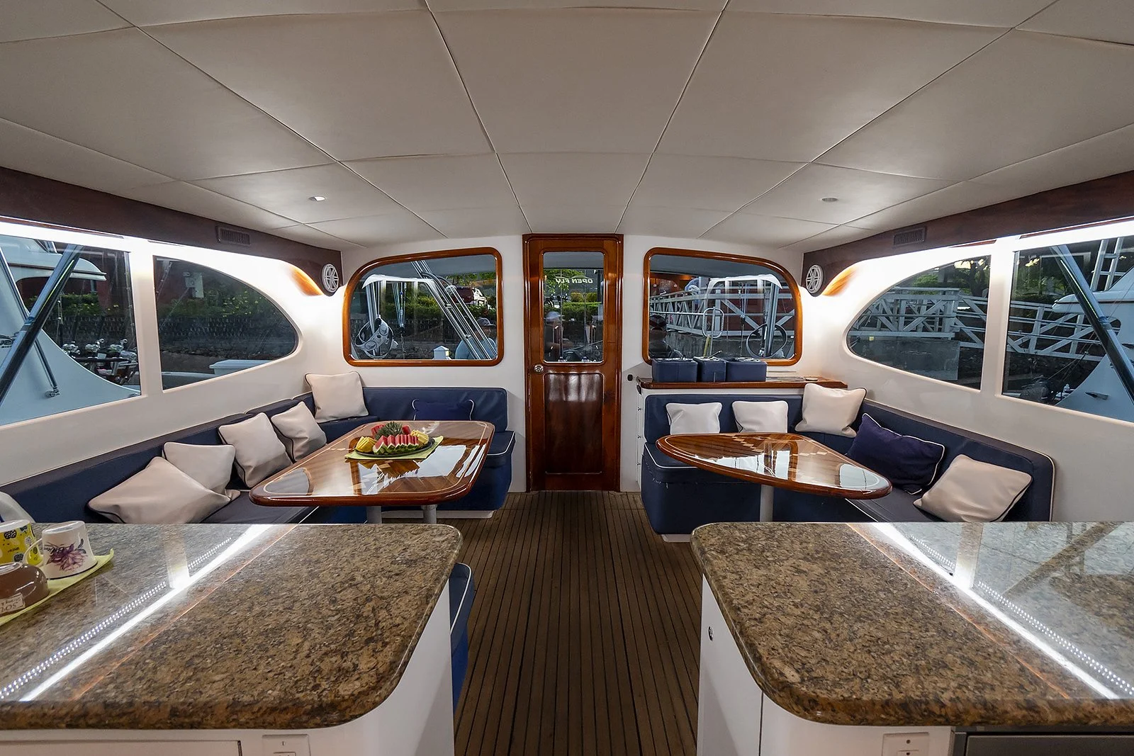 Interior of a boat with seating, tables, and large windows showing outside boat dock and water.