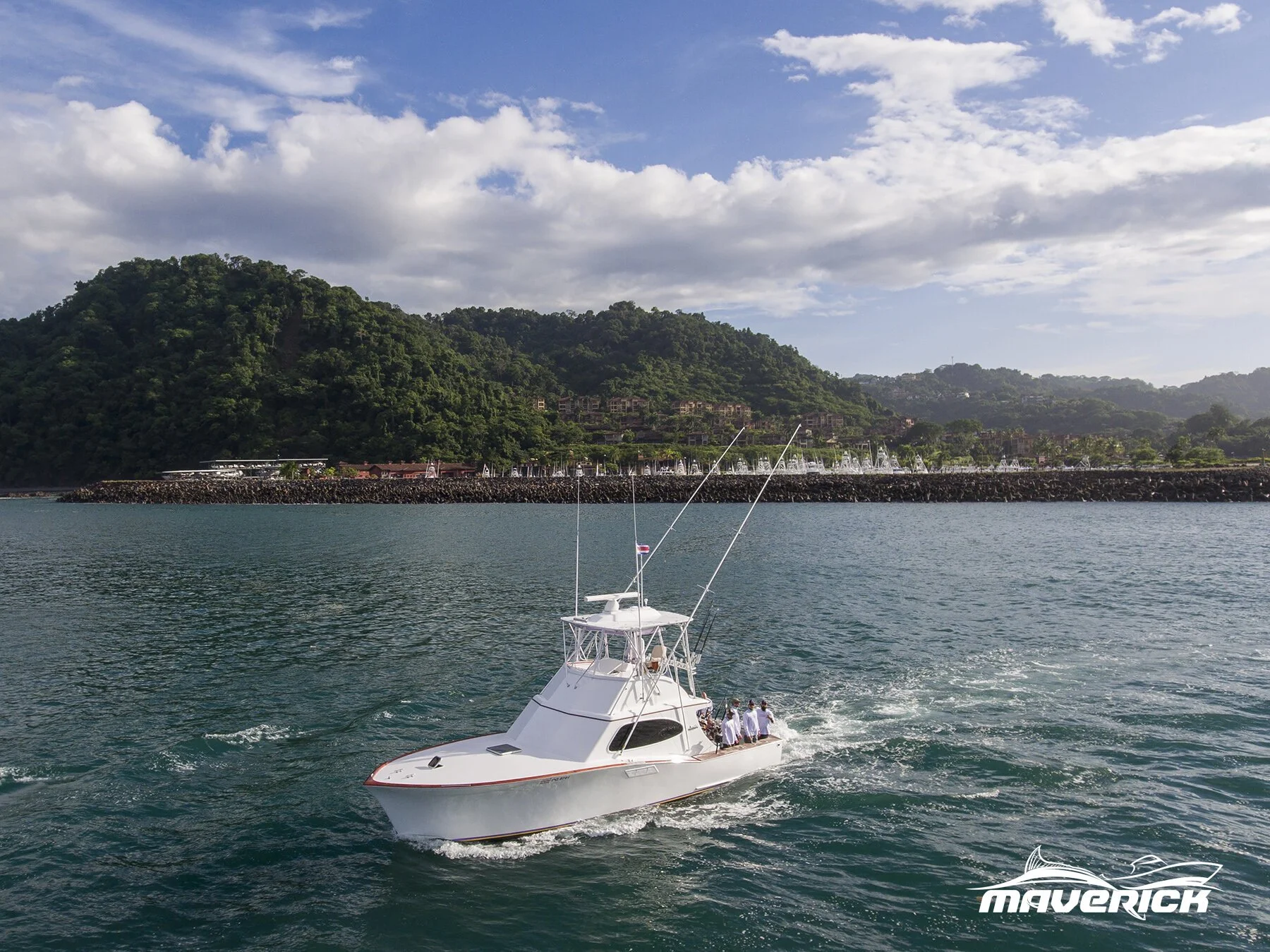 A white Maverick boat with three people aboard sailing in calm waters near a green, hilly coastline with buildings and a marina in the background, under a partly cloudy sky.