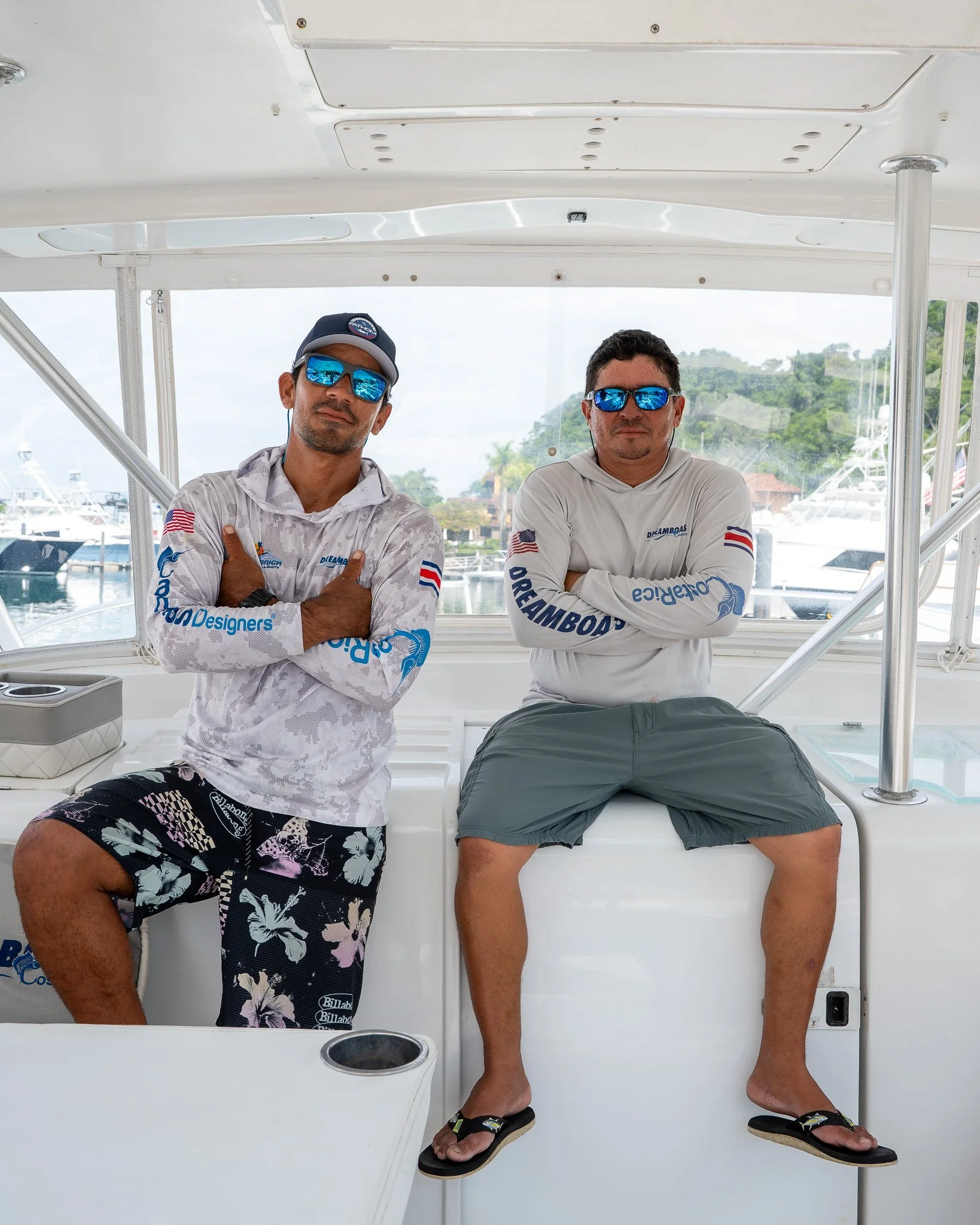 Two men sitting on a boat with a marina and boats in the background, both wearing sunglasses and casual summer clothing with branding.