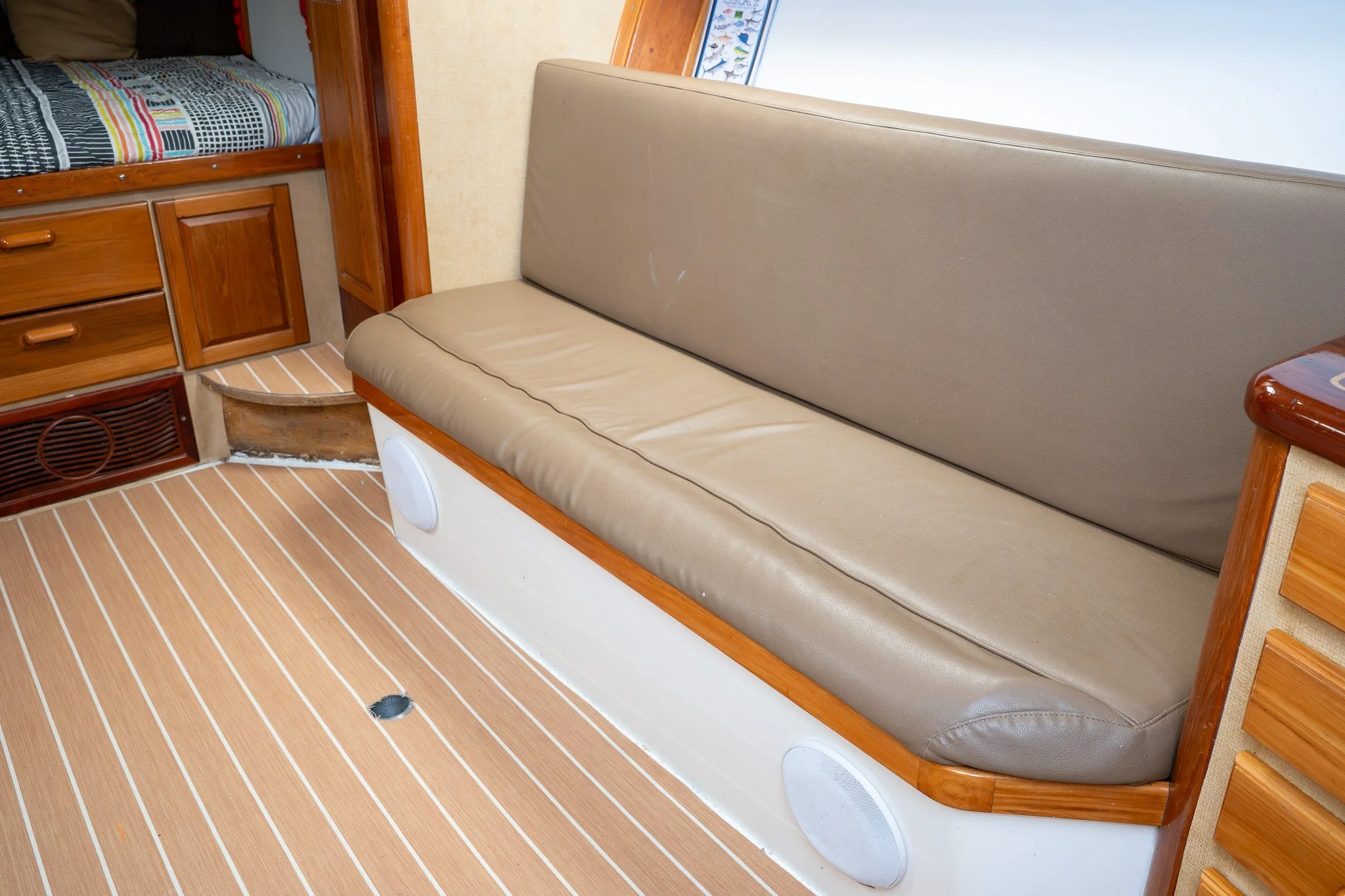 Interior of a boat or RV showing a beige bench with leather upholstery, wood trim, built-in speaker, and a striped tan carpet with a hole. A small bed with a colorful striped blanket is visible in the background.