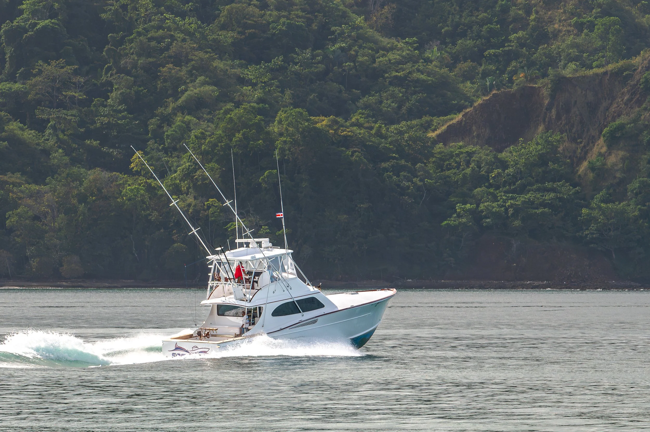 A white motor yacht speeding across a body of water with a lush, green forested hillside in the background.