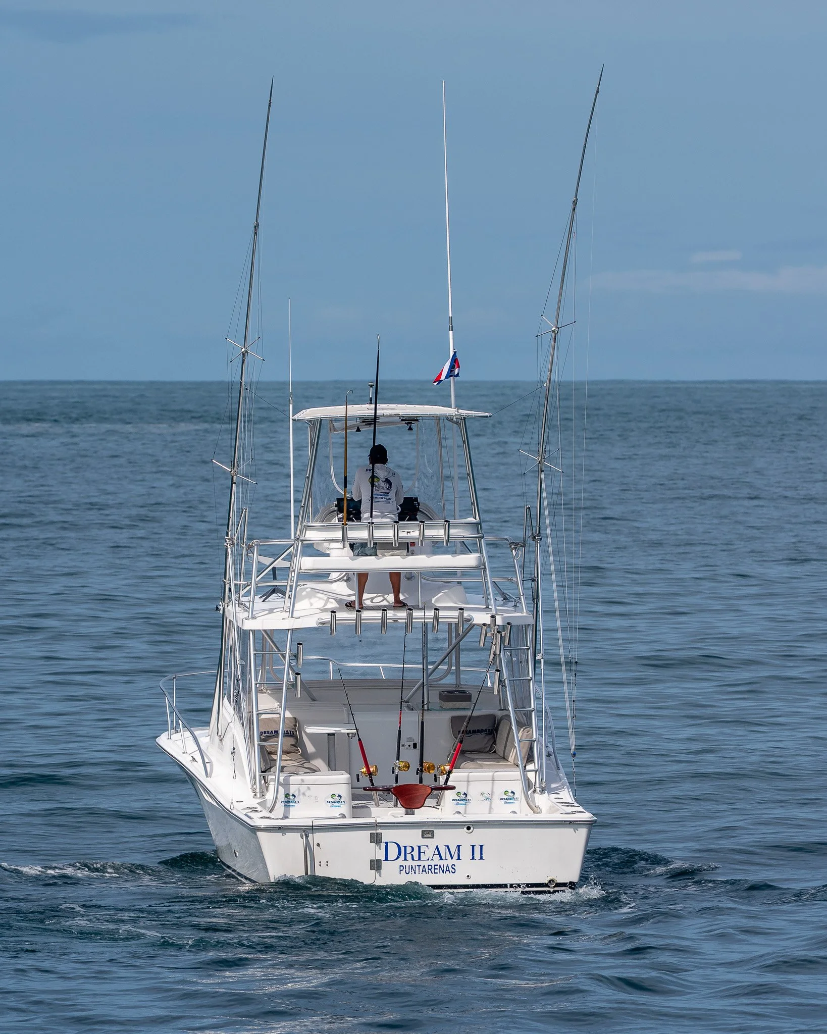 A white fishing boat named 'Dream II' cruising on the ocean, with a person standing on the upper deck operating the boat, under a blue sky.