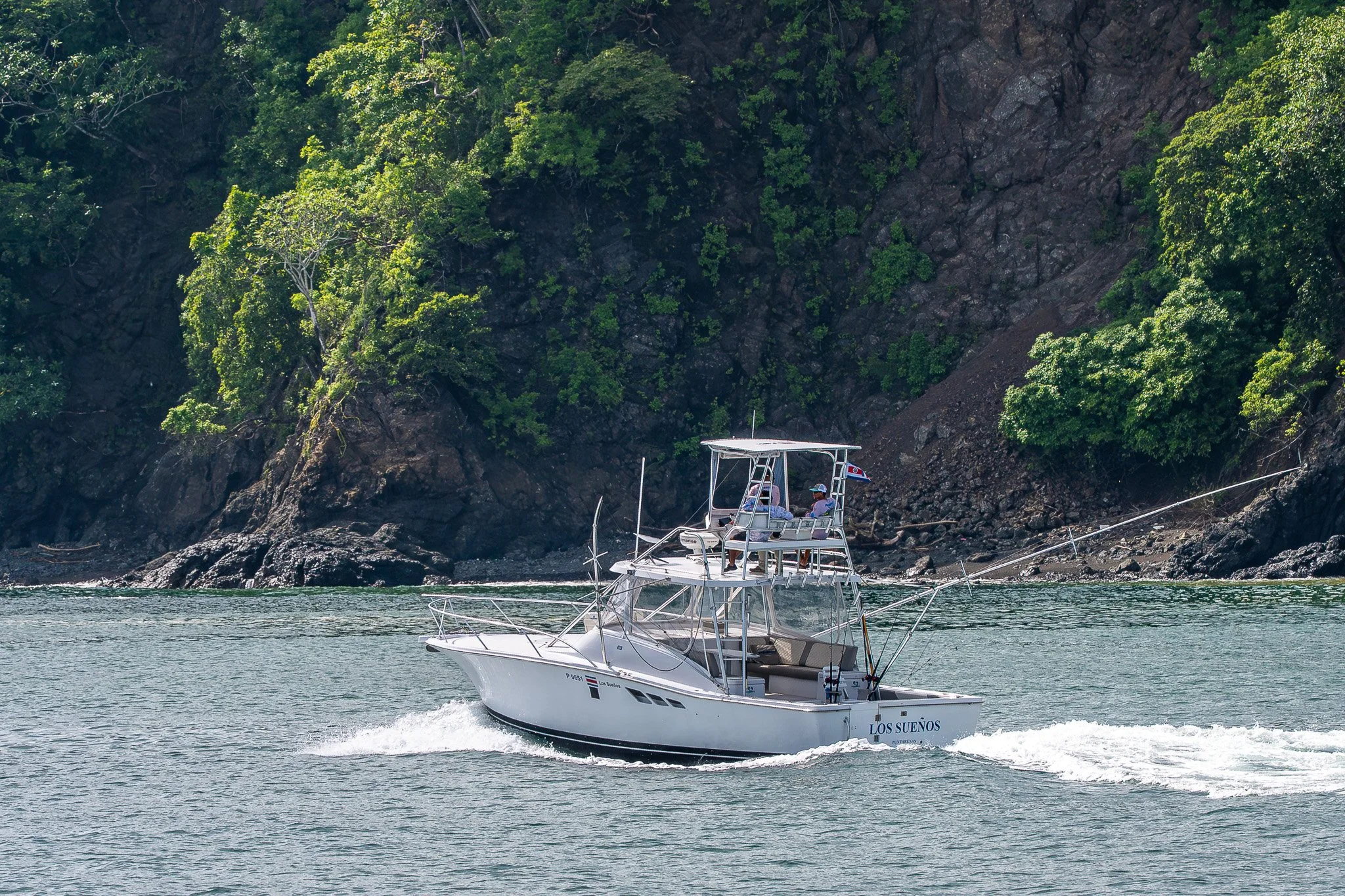 A white boat named 'Los Sueños' sailing on water with green lush cliffs in the background.