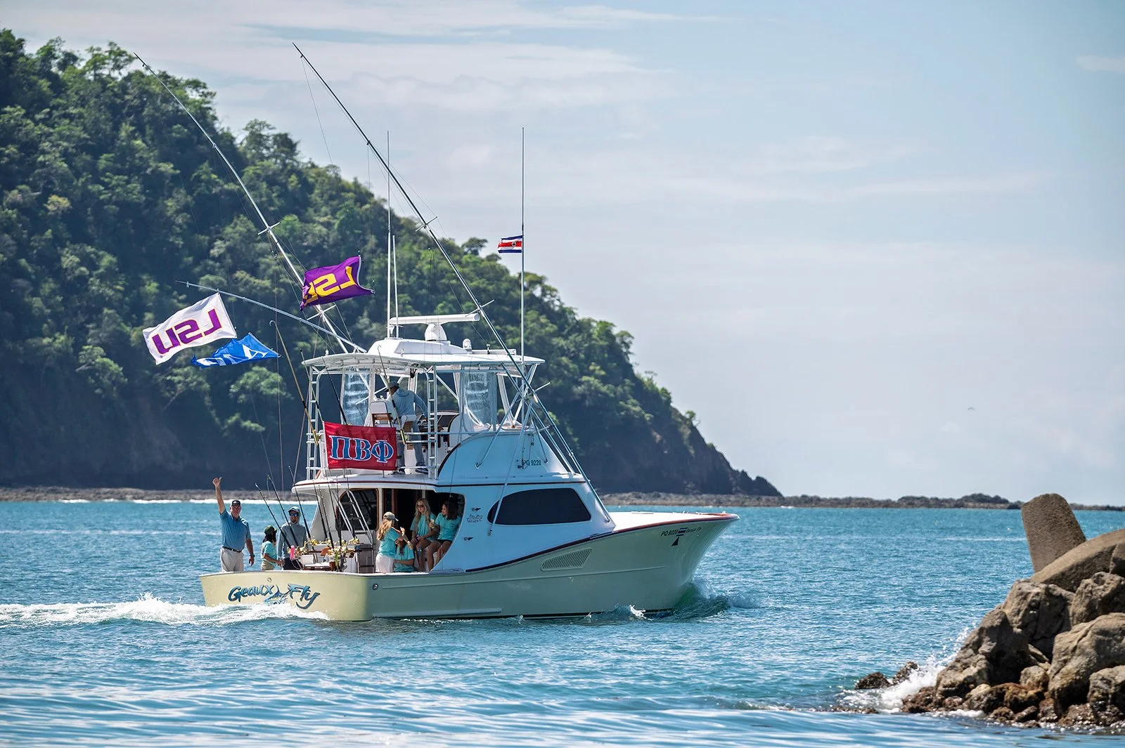 A yacht with people on board, flying flags, moving through turquoise water near a rocky shoreline with a green, forested hill in the background.