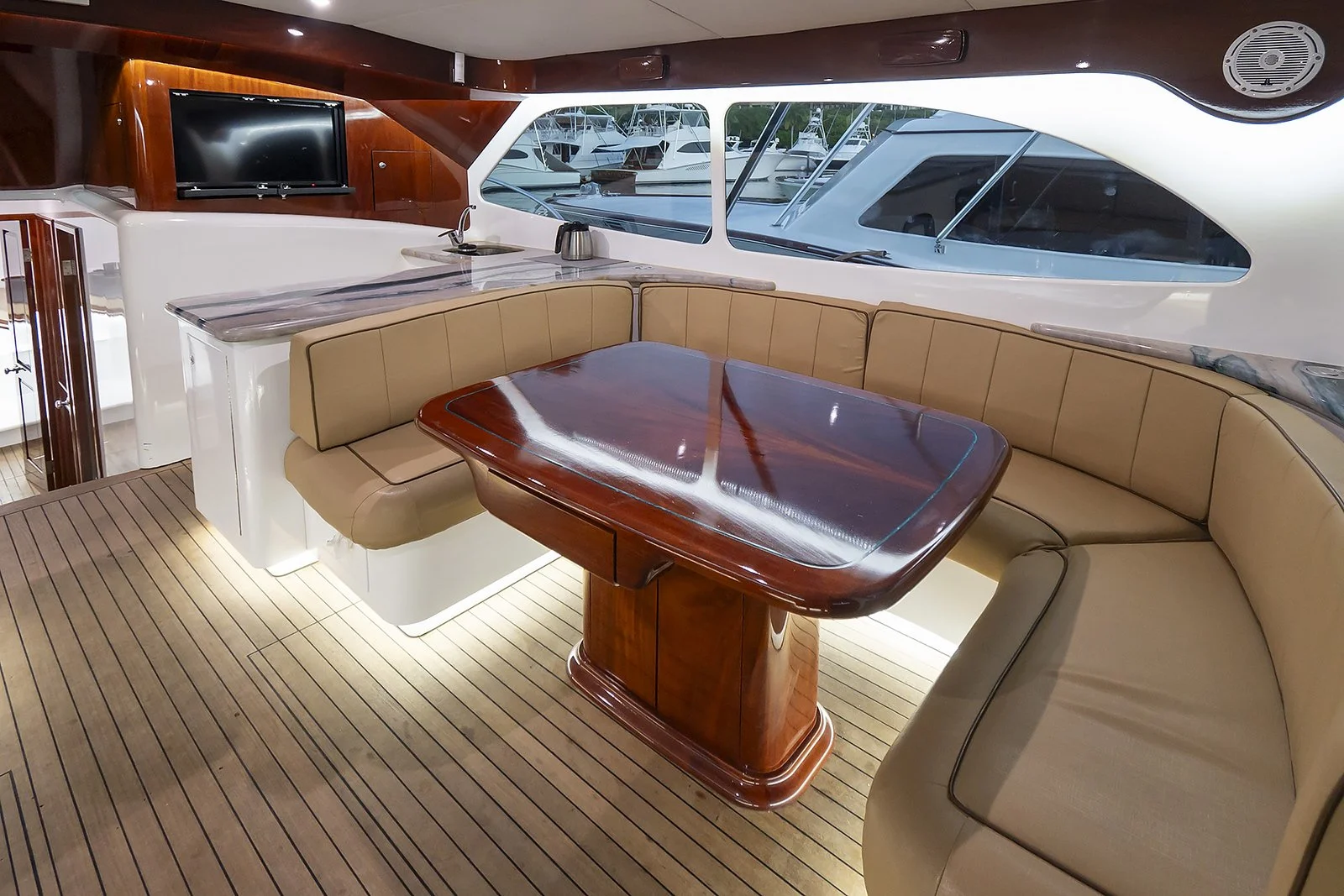 Interior of a yacht featuring a U-shaped beige leather sofa, a polished wooden table, a small kitchenette with a sink, and large windows overlooking moored boats in a marina.