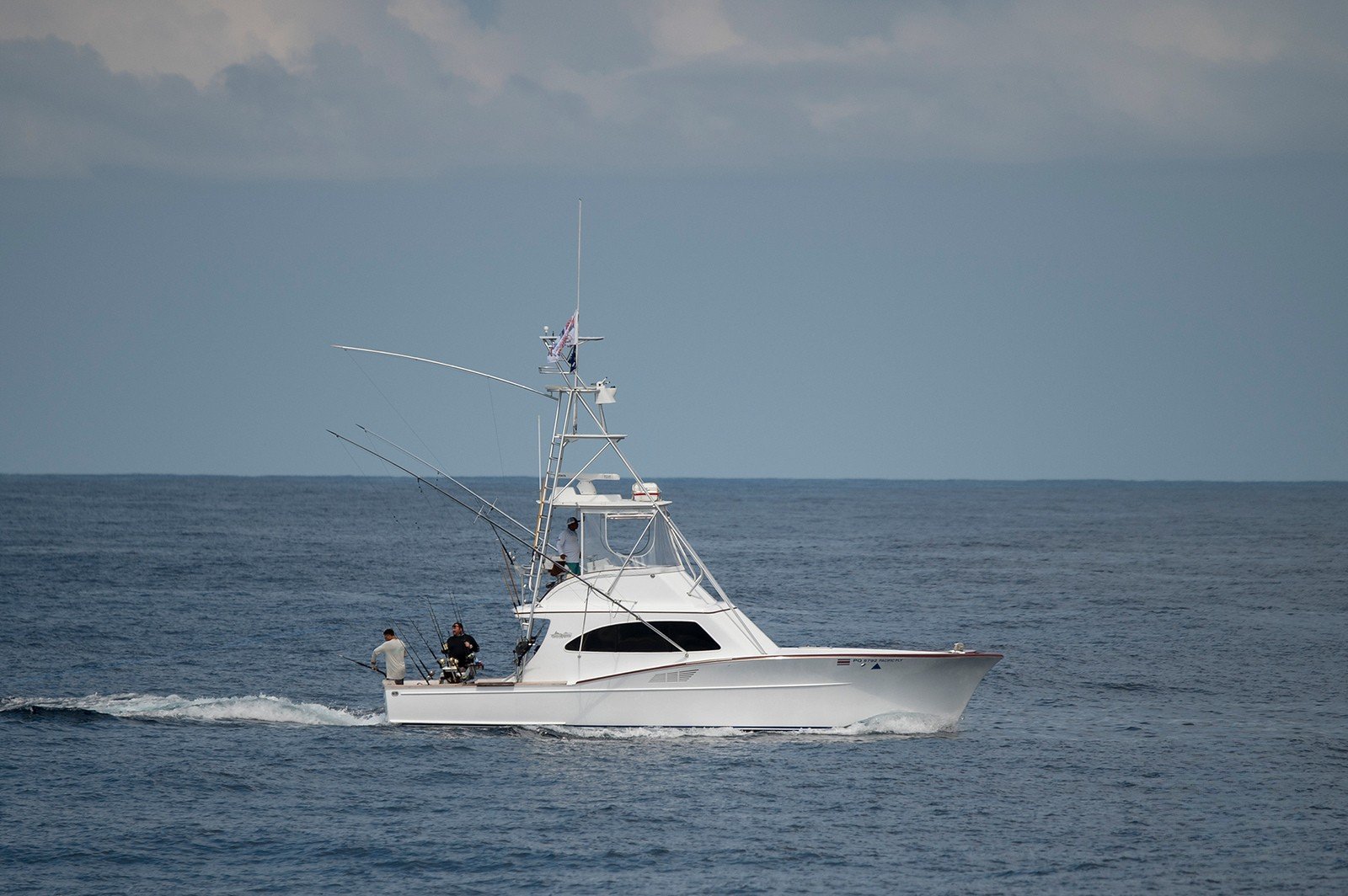 A white fishing boat with four people on board sailing in the ocean with a cloudy sky overhead.