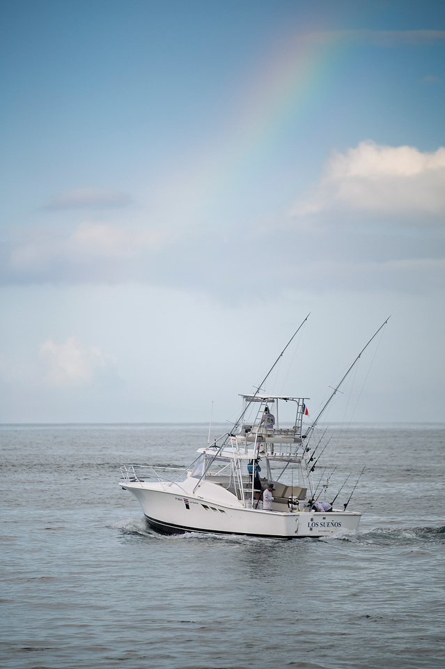 A white fishing boat named Los Sueños moving on calm water under a partly cloudy sky with a faint rainbow.
