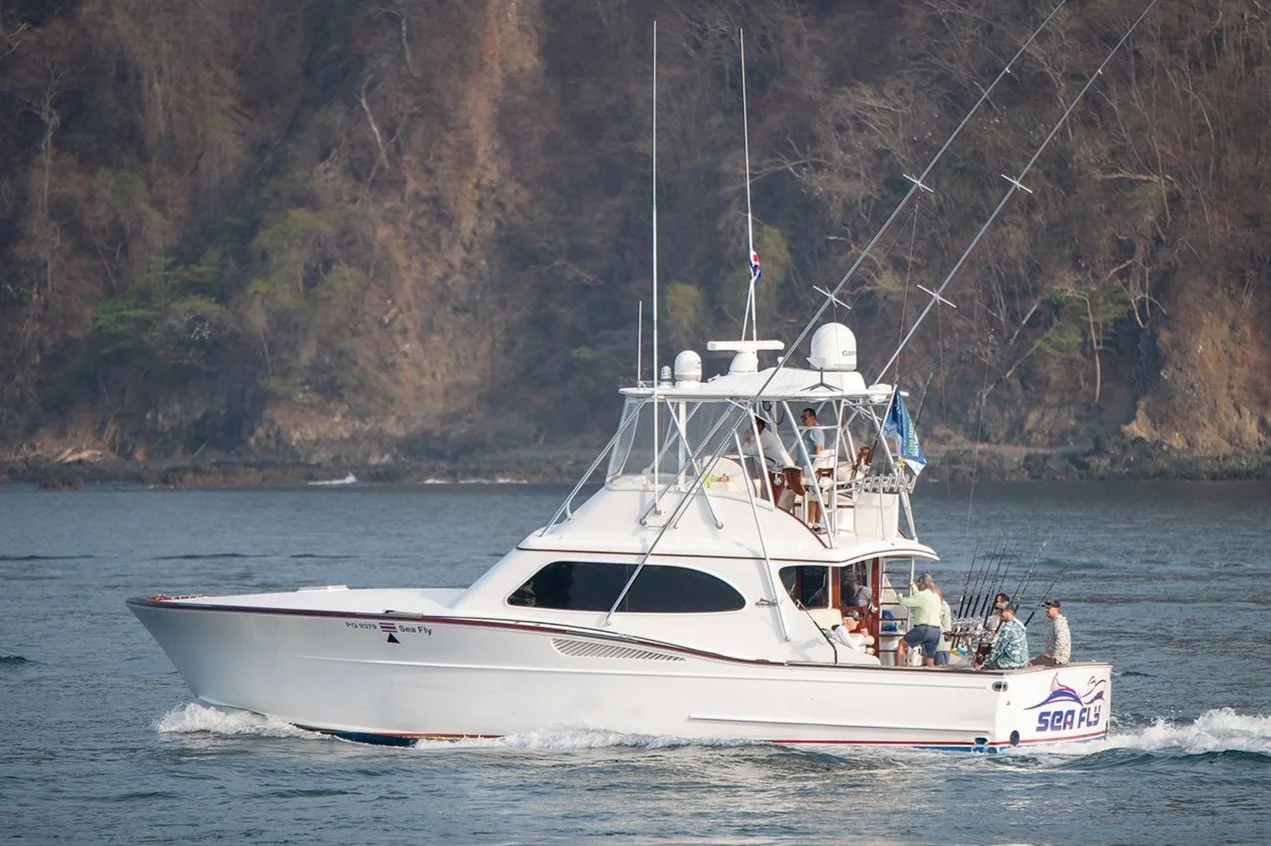 A white yacht with the words 'Sea Fly' on the side, sailing on calm water near a rocky, wooded shoreline, with people on deck fishing and relaxing.