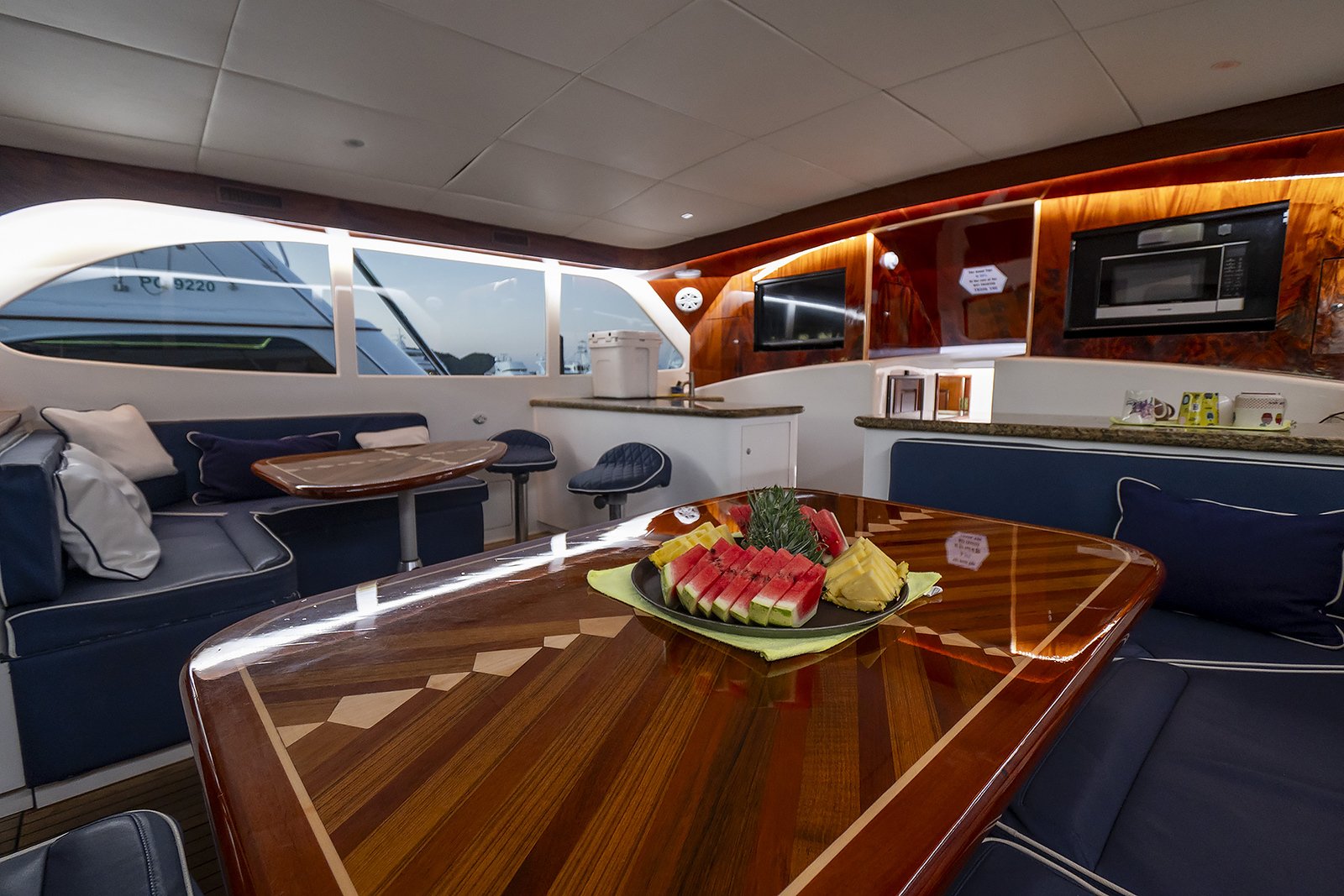 Interior of a luxury yacht with a wooden table topped with sliced fruit including watermelon, pineapple, and mango, set in a stylish lounge area with navy blue cushions, a window view of the water and sky outside.