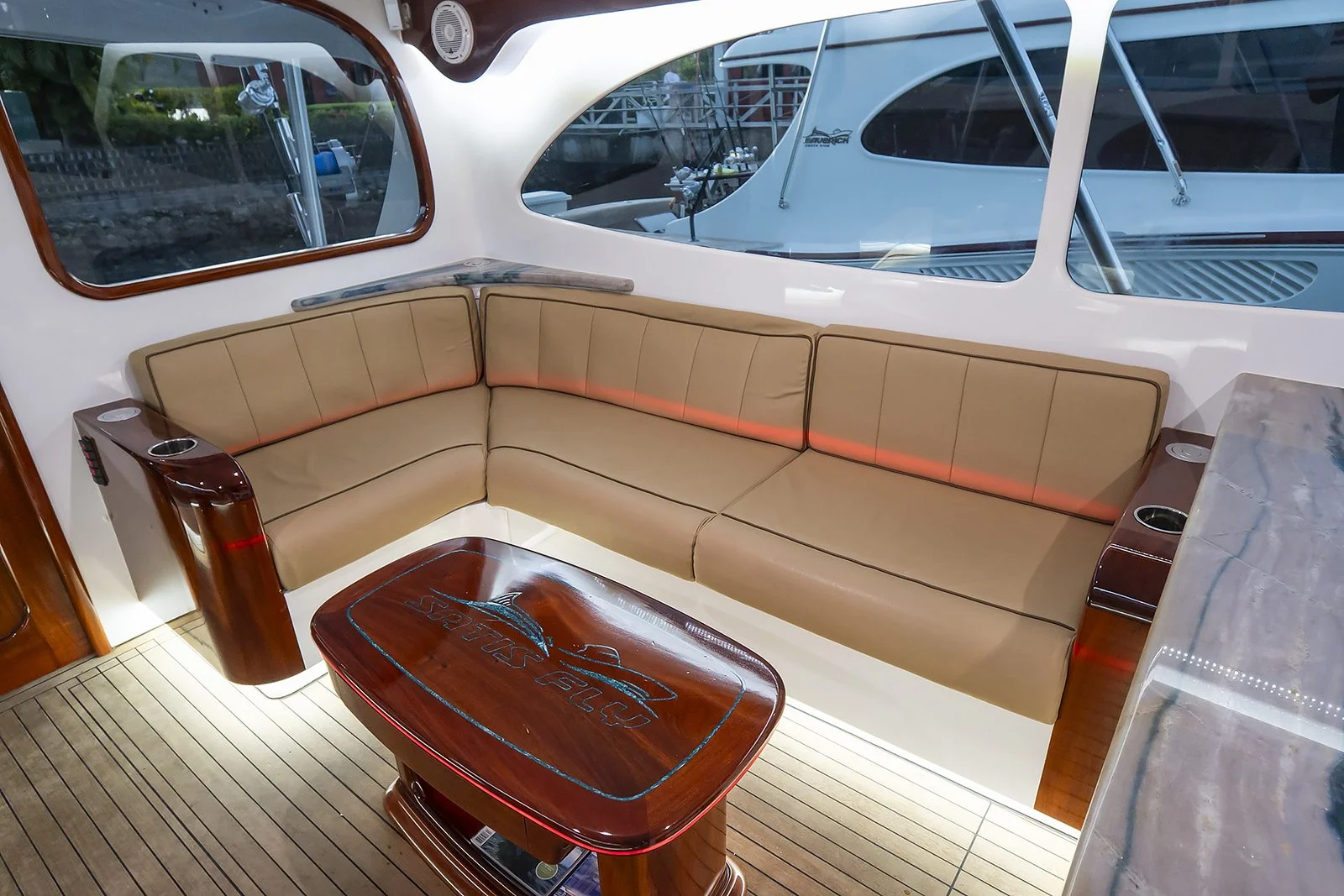 Interior of a yacht with a tan leather sofa, wooden table, and large windows showing masts and boats outside.