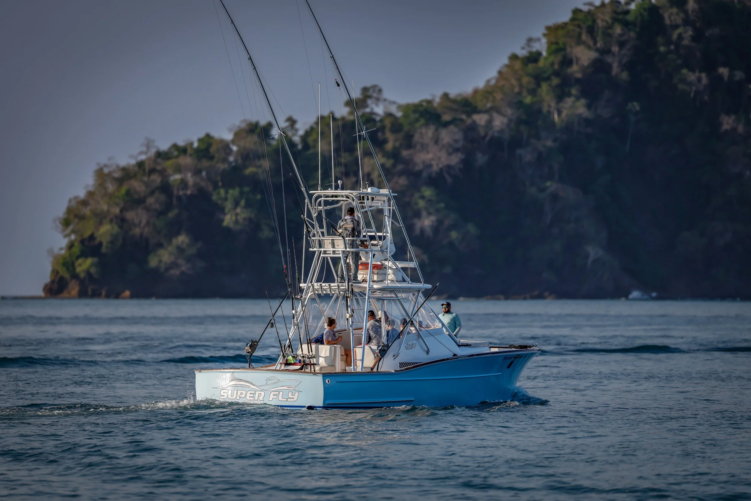 A blue and white fishing yacht named 'Super Fly' cruising on the ocean with four people onboard, near a lush, green island in the background.