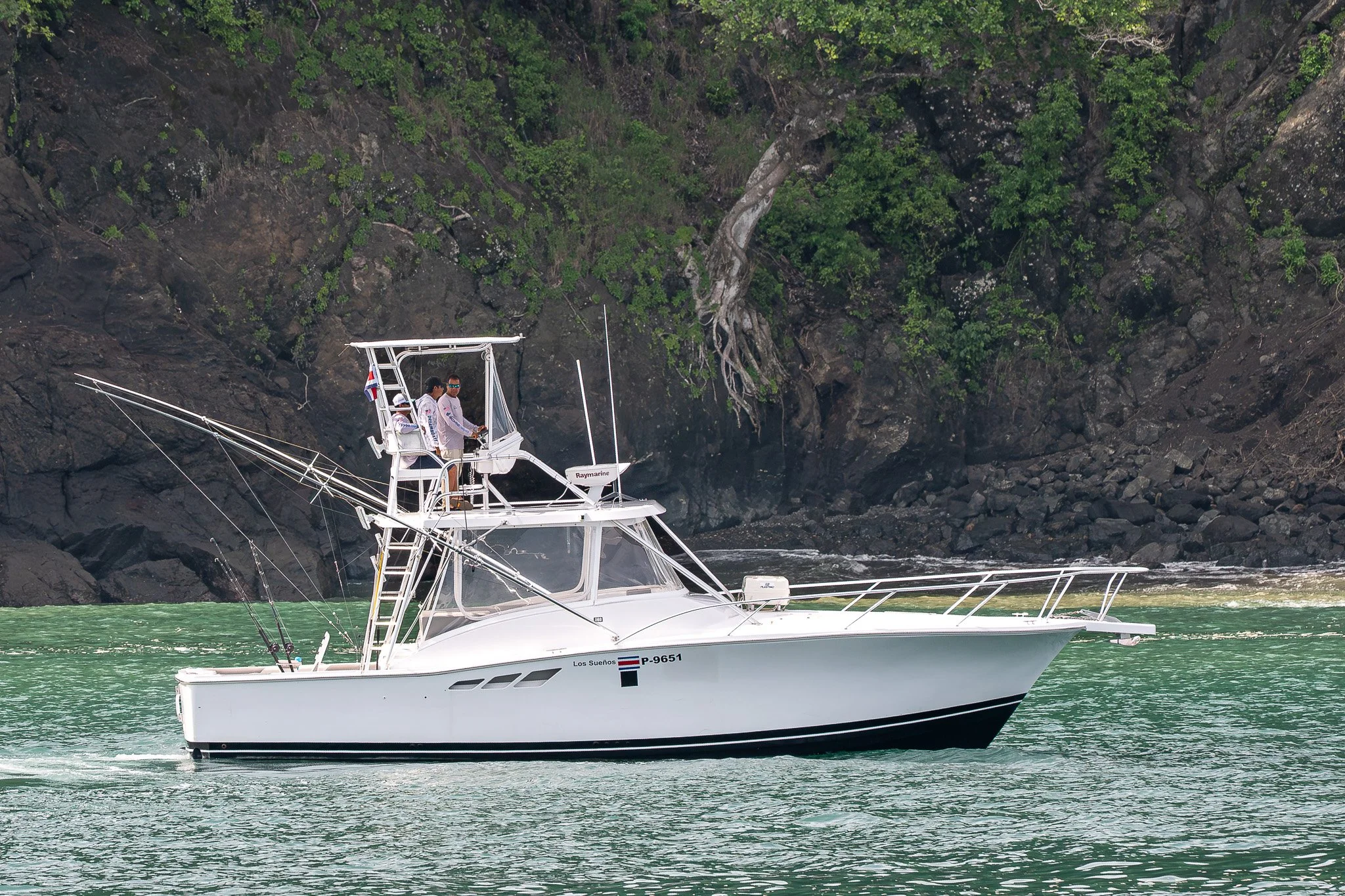 A white fishing boat with people on the upper deck sailing in a turquoise body of water near a rocky shoreline with green trees.