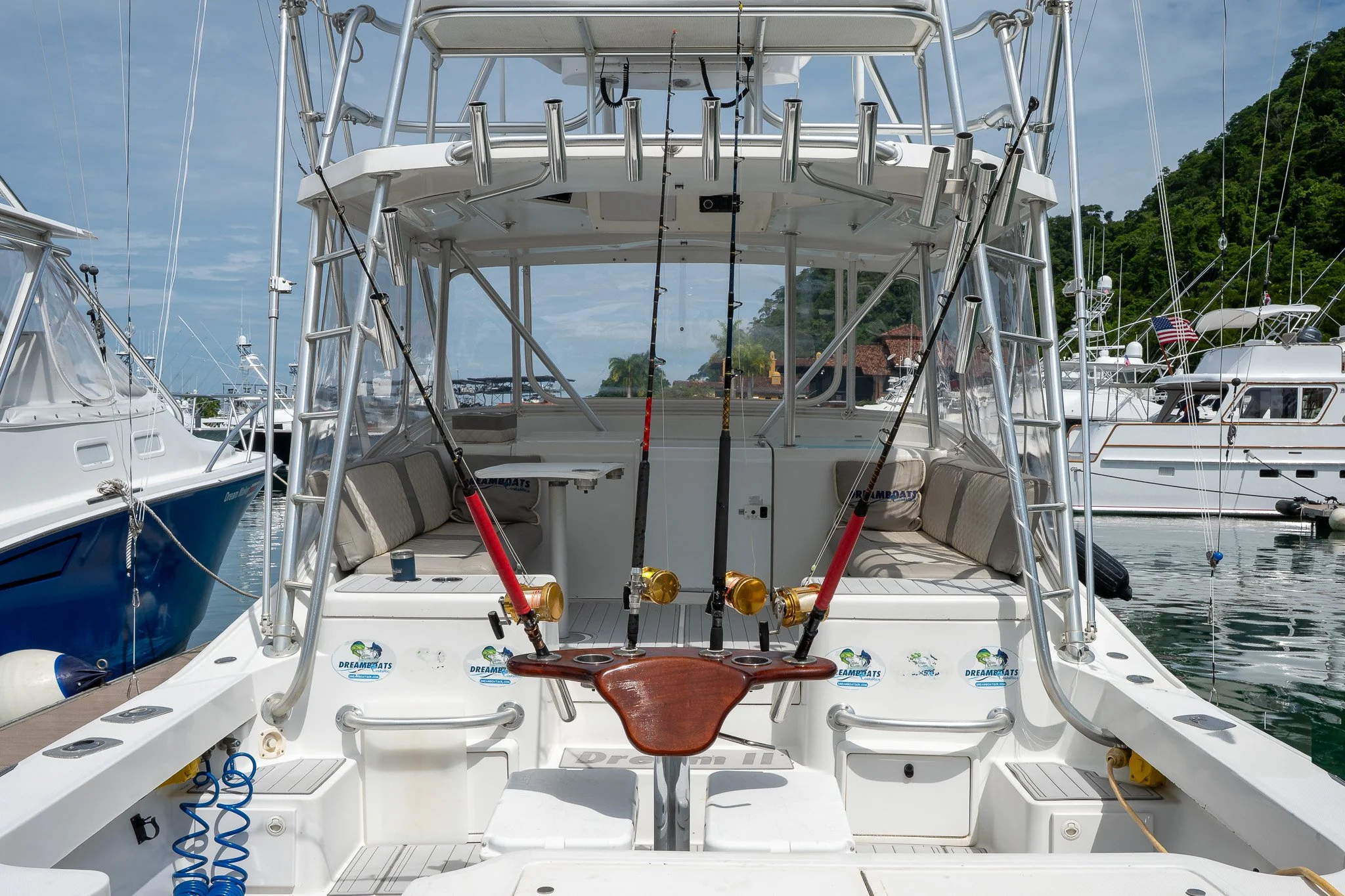 Rear view of a fishing boat docked at a marina, with fishing rods set up on the boat and other boats visible in the background.