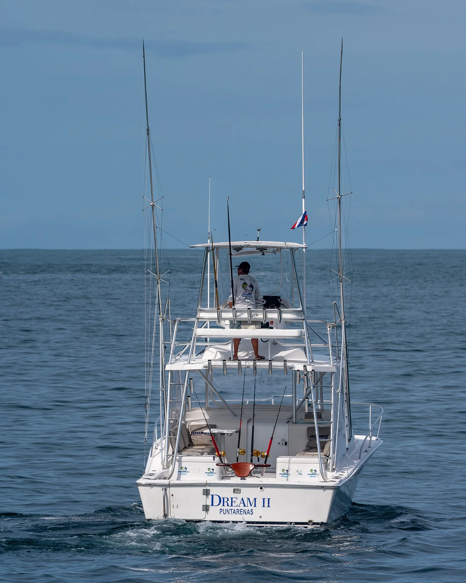 A white fishing boat named Dream II with two fishing rods on the back, floating on calm water with a person standing on the upper deck.