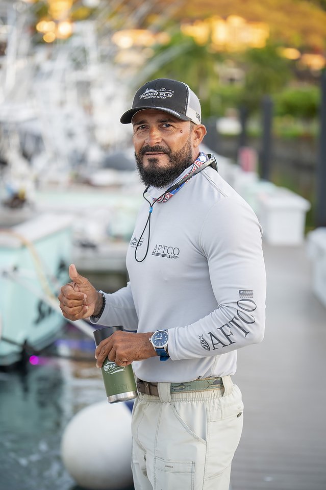 A man with a beard and wearing a baseball cap, long sleeve shirt, and beige pants stands near boats at a marina, giving a thumbs-up and holding a green travel mug.