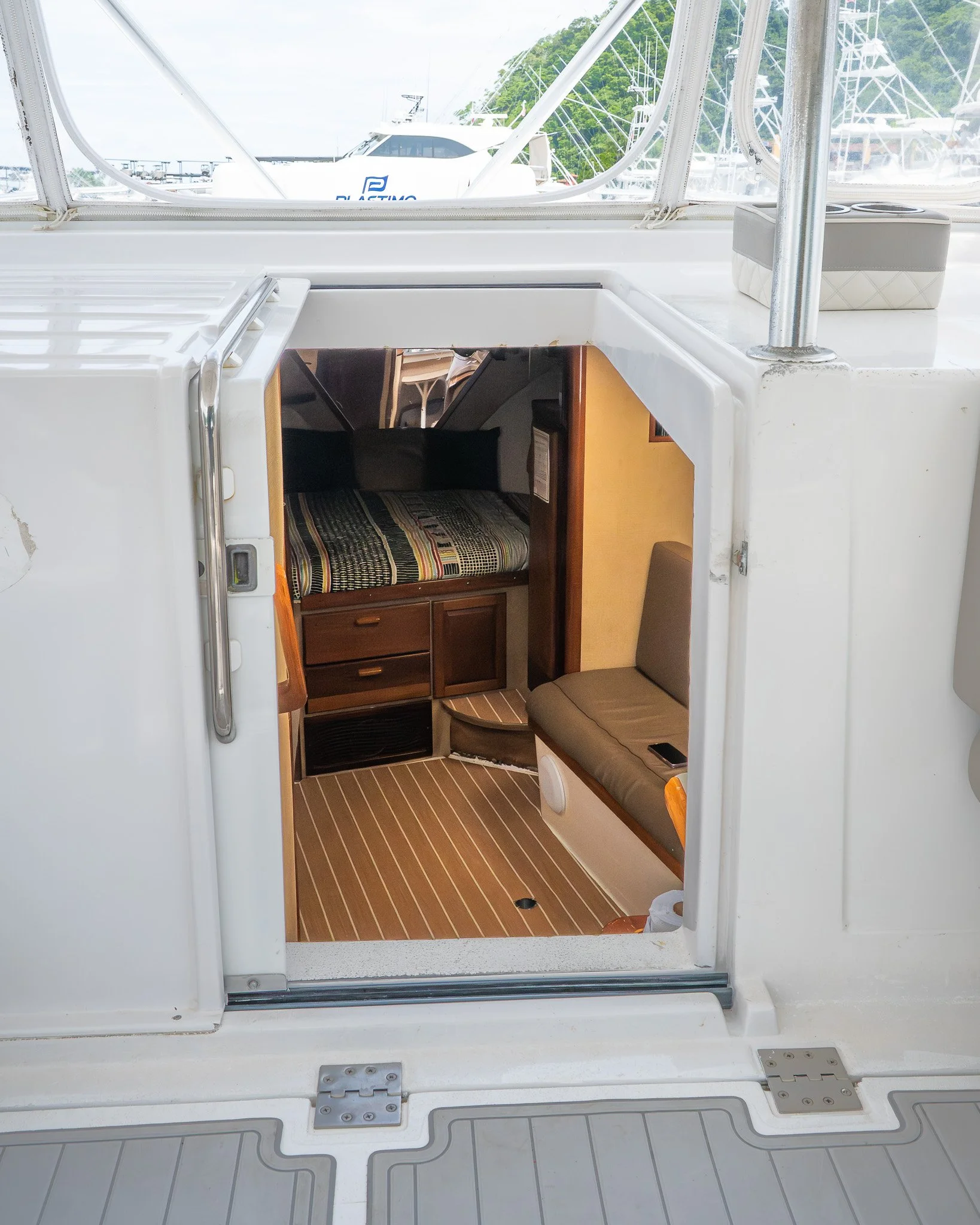 Interior of a boat cabin with a bed, sitting area, and wooden cabinets, viewed through an open hatch, with boats and marina visible outside.