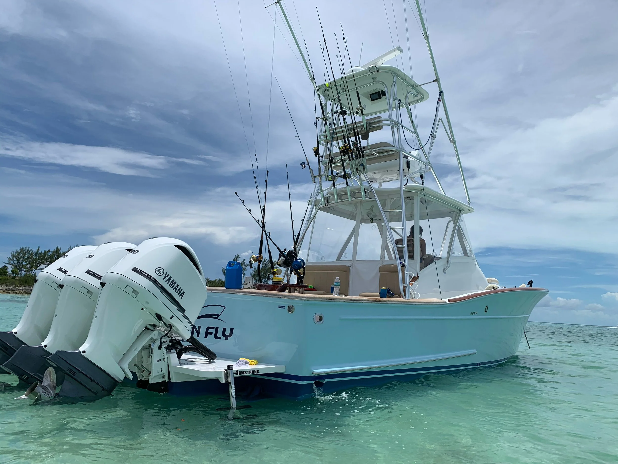 A white fishing boat with three Yamaha outboard motors, equipped with multiple fishing rods, is anchored in clear, shallow water under a partly cloudy sky.