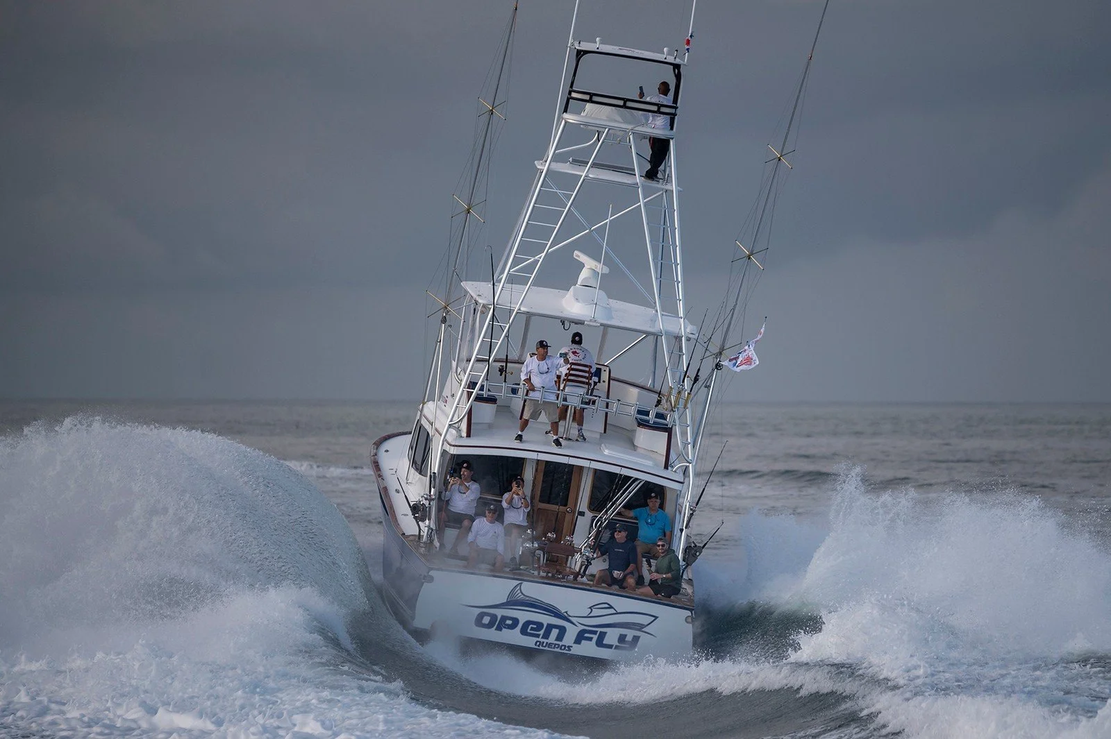 A group of people on a white fishing boat named 'Open Fly' speeding through the water, creating waves and spray, with a cloudy sky in the background.