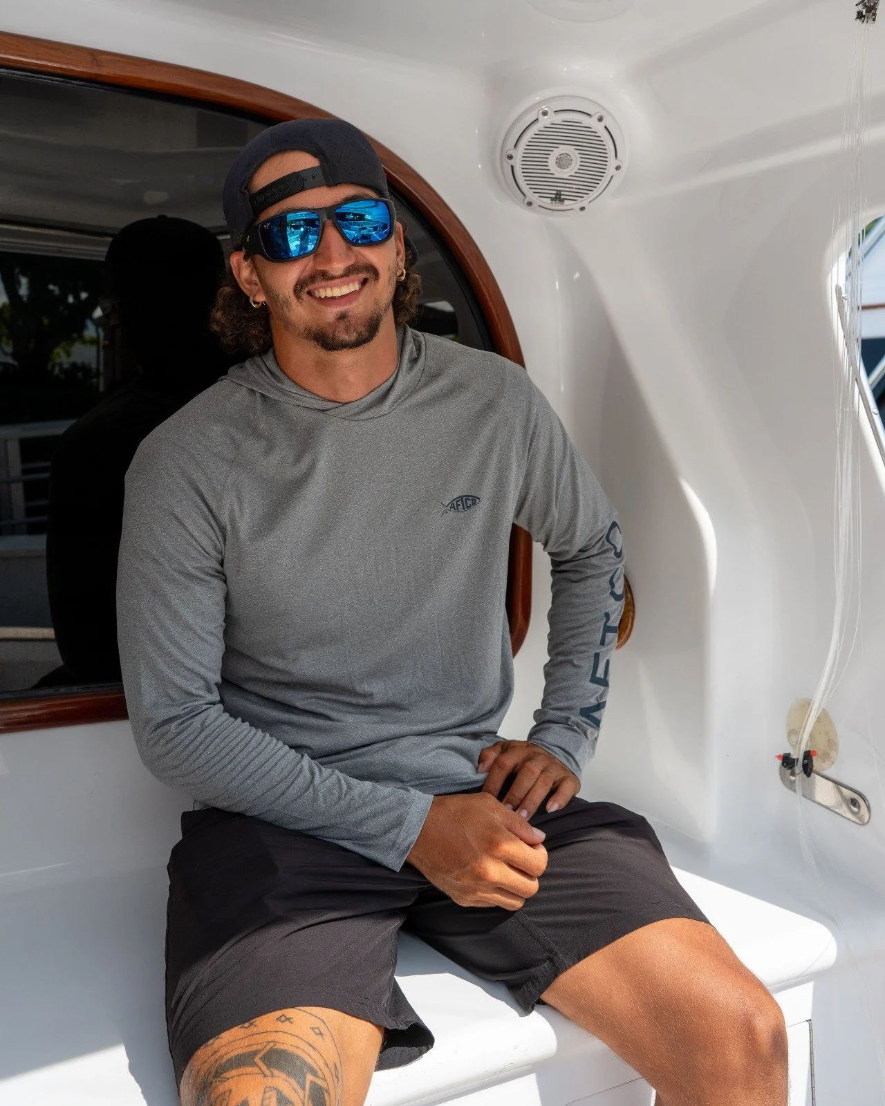 Young man sitting on a boat with sunglasses, gray long sleeve shirt, black shorts, and a tattoo on his thigh, smiling.