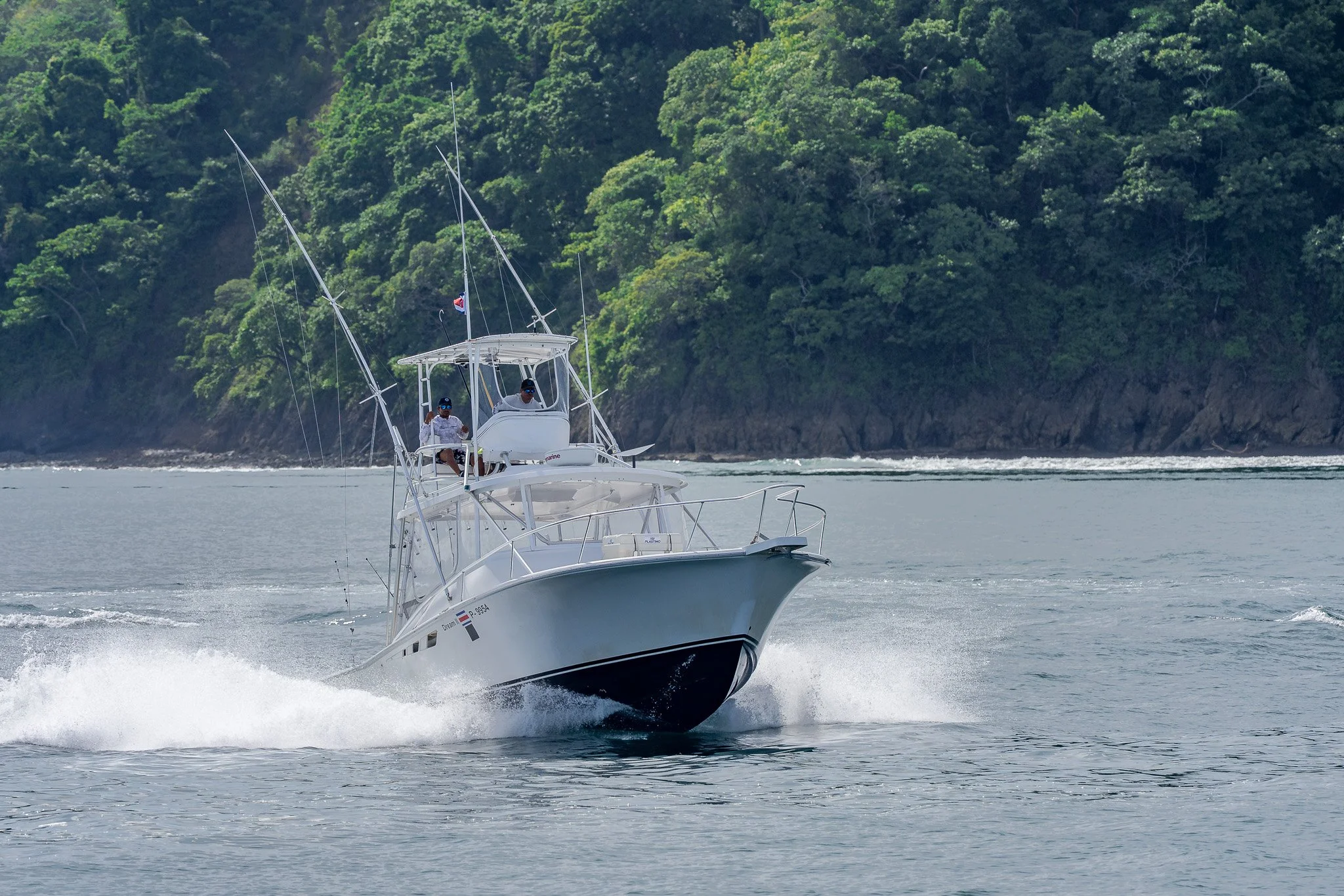 A white motor yacht with two men onboard, sailing on calm water near a lush green forested shoreline.