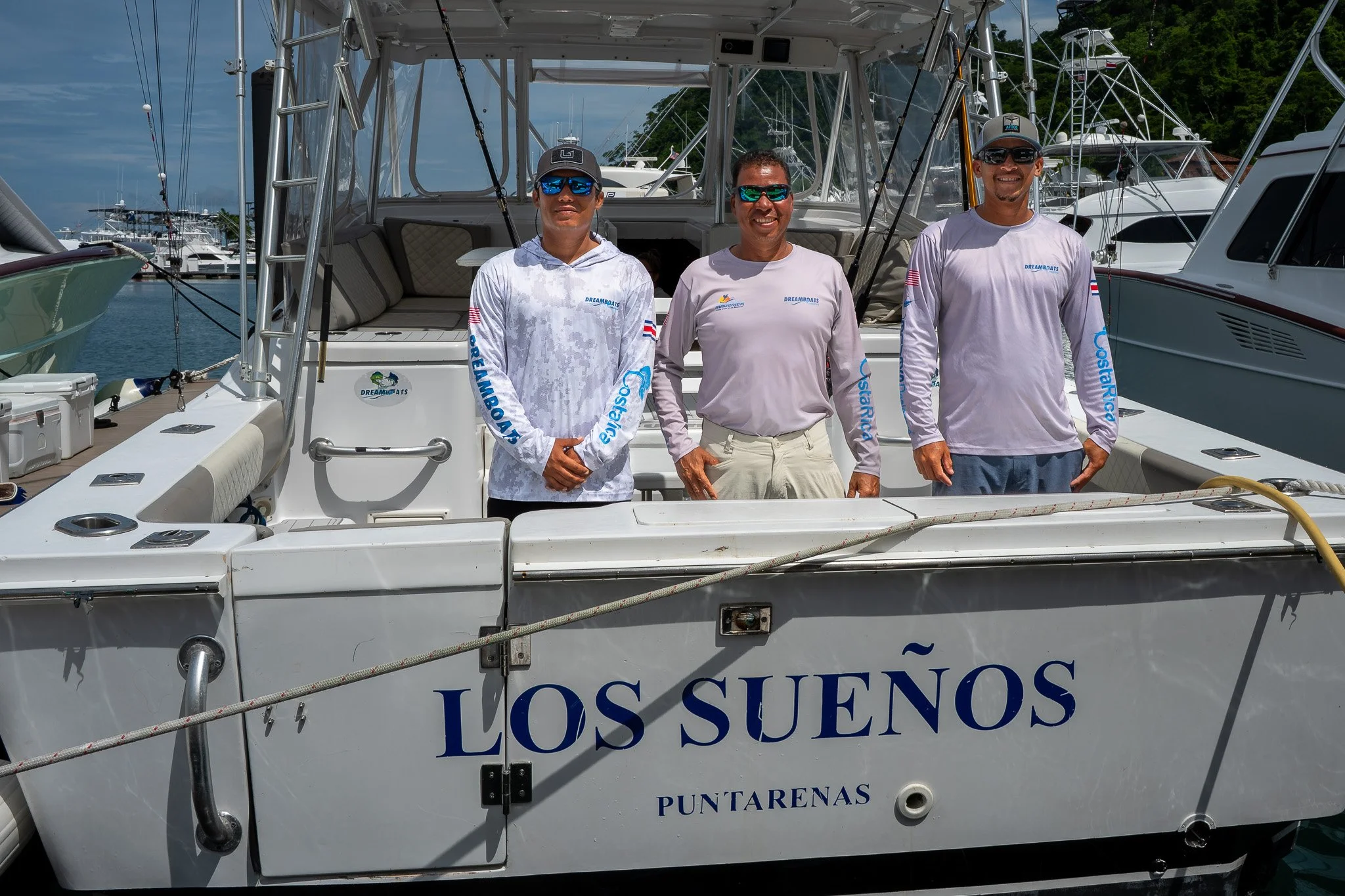 Three men standing on a boat named 'Los Sueños' in a marina, smiling and wearing casual shirts and sunglasses.