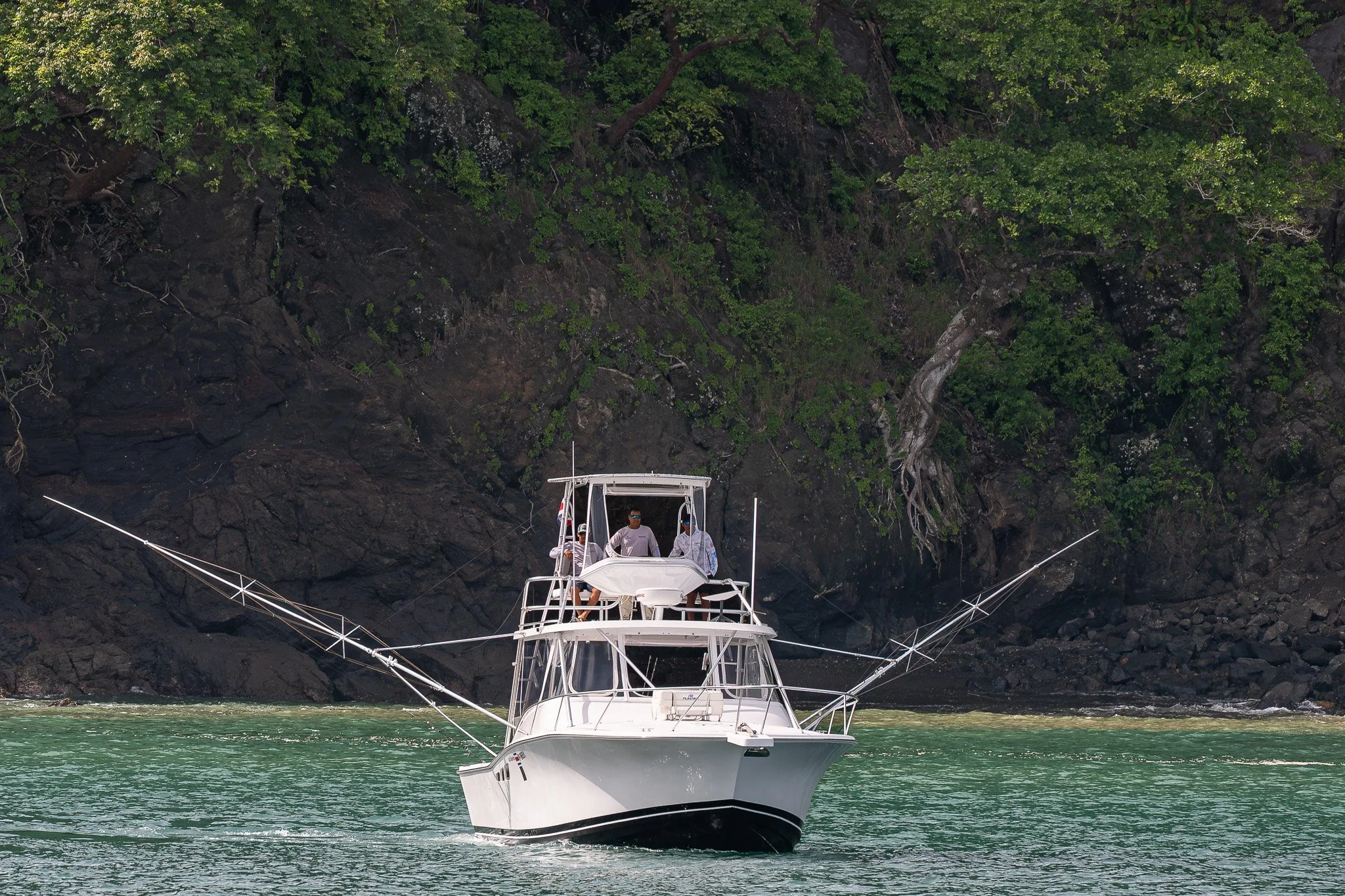 A white fishing boat with people on the upper deck, anchored near a rocky shoreline with dense green trees.