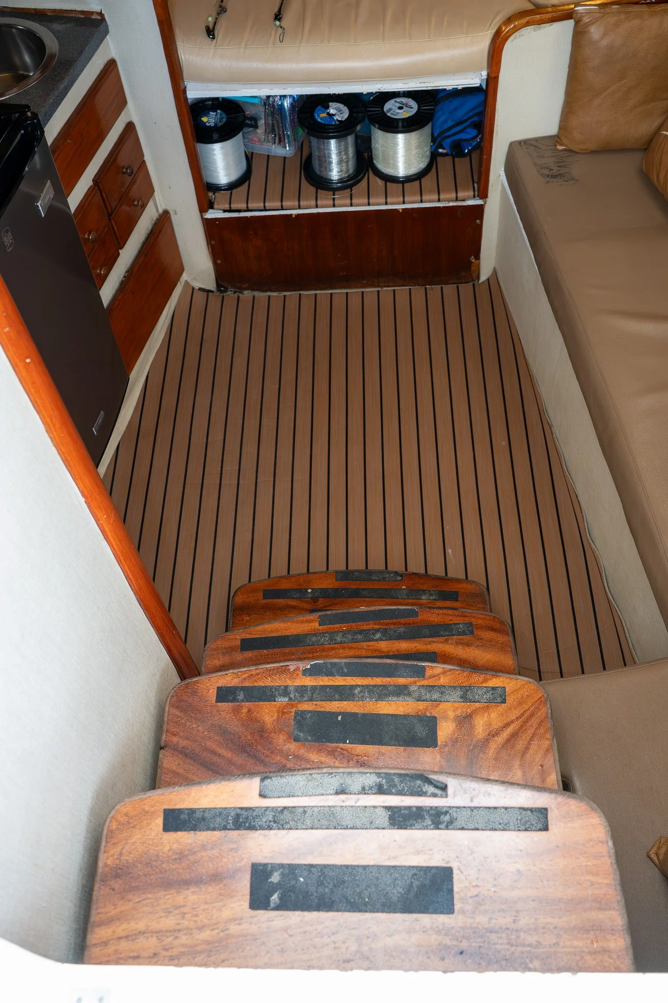 A small interior space with a staggered set of wooden stairs, a beige sofa, a wooden floor with narrow planks, and a kitchen area with drawers, a small refrigerator, and a shelf with canned goods.