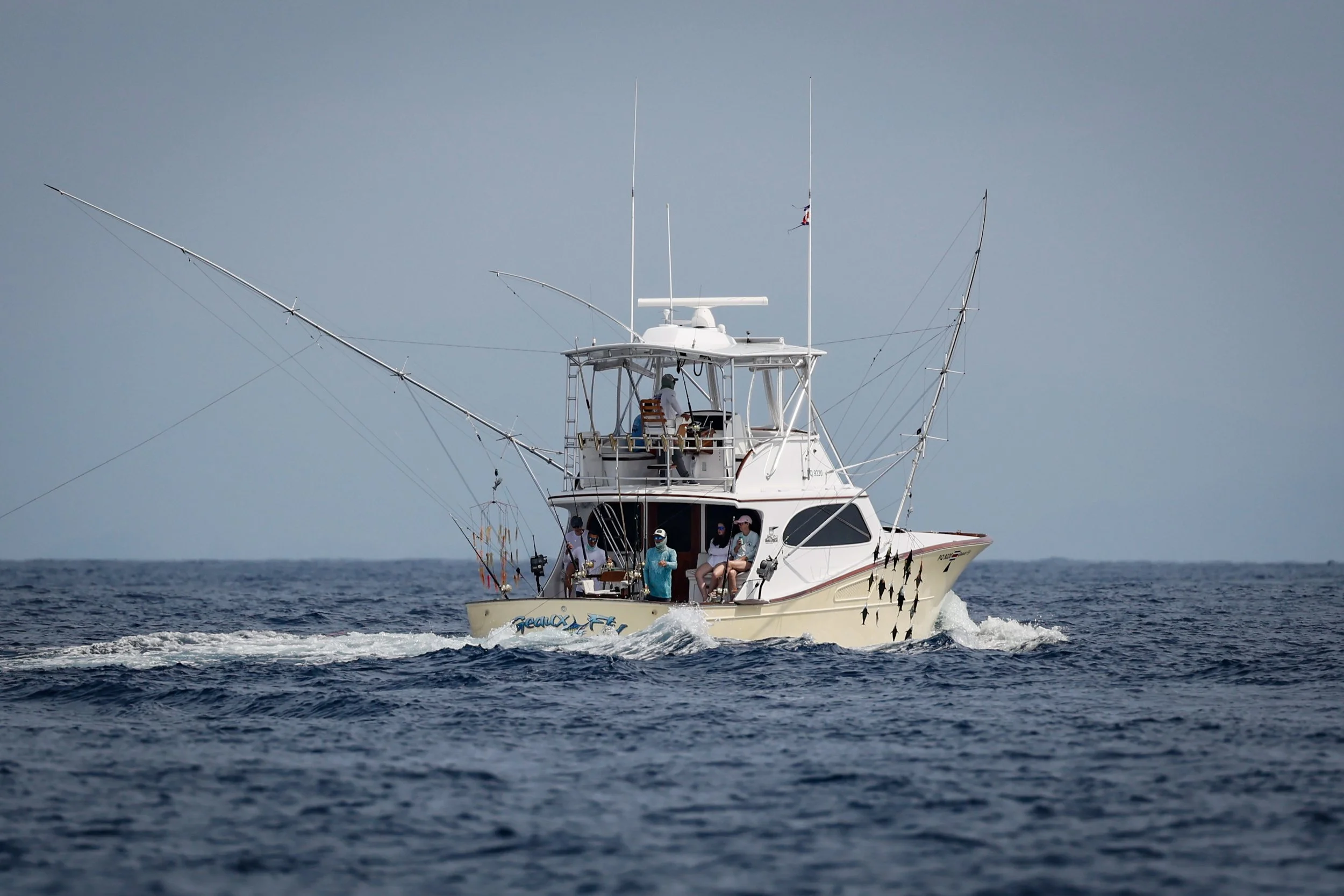 A white fishing boat with multiple fishing rods underway on the water, with several people on deck, some sitting and some standing, as it moves through the ocean.