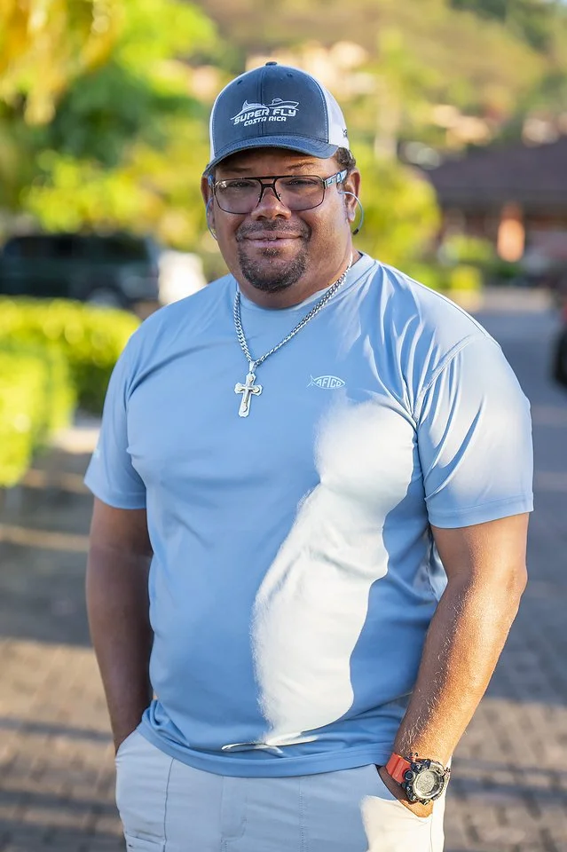 A man standing outdoors on a sunny day, wearing glasses, a baseball cap, a light blue athletic shirt, a cross necklace, and a wristwatch. He is smiling with his hands in his pockets.
