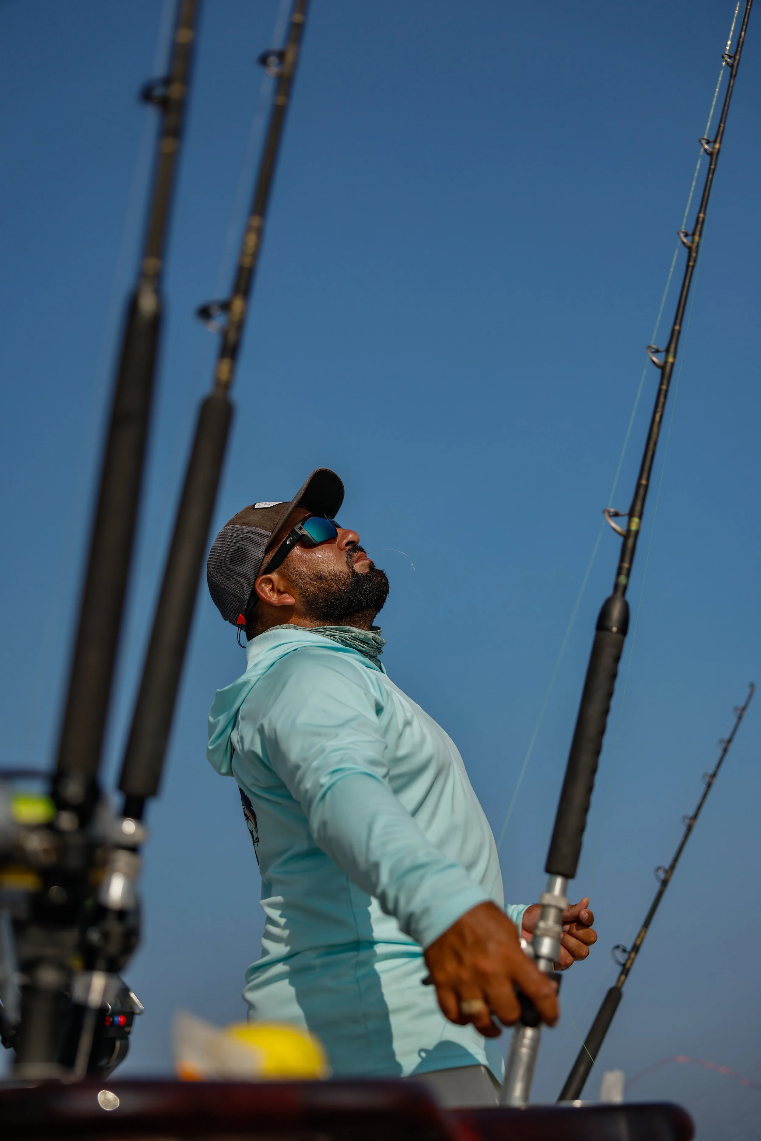 A man in sunglasses, a cap, and a light blue fishing shirt is holding fishing rods on a boat, looking up at the sky.