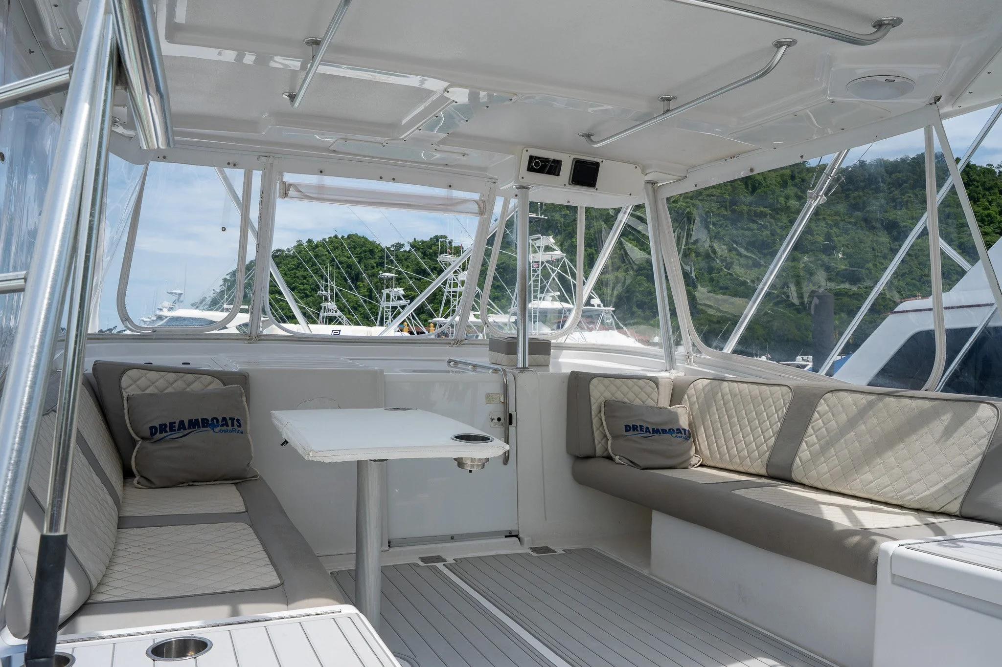 Interior of a yacht with cushioned seating, a small table, pillows with "DREAMBOATS Coastal" logo, large windows, and a view of other boats and green hills outside.