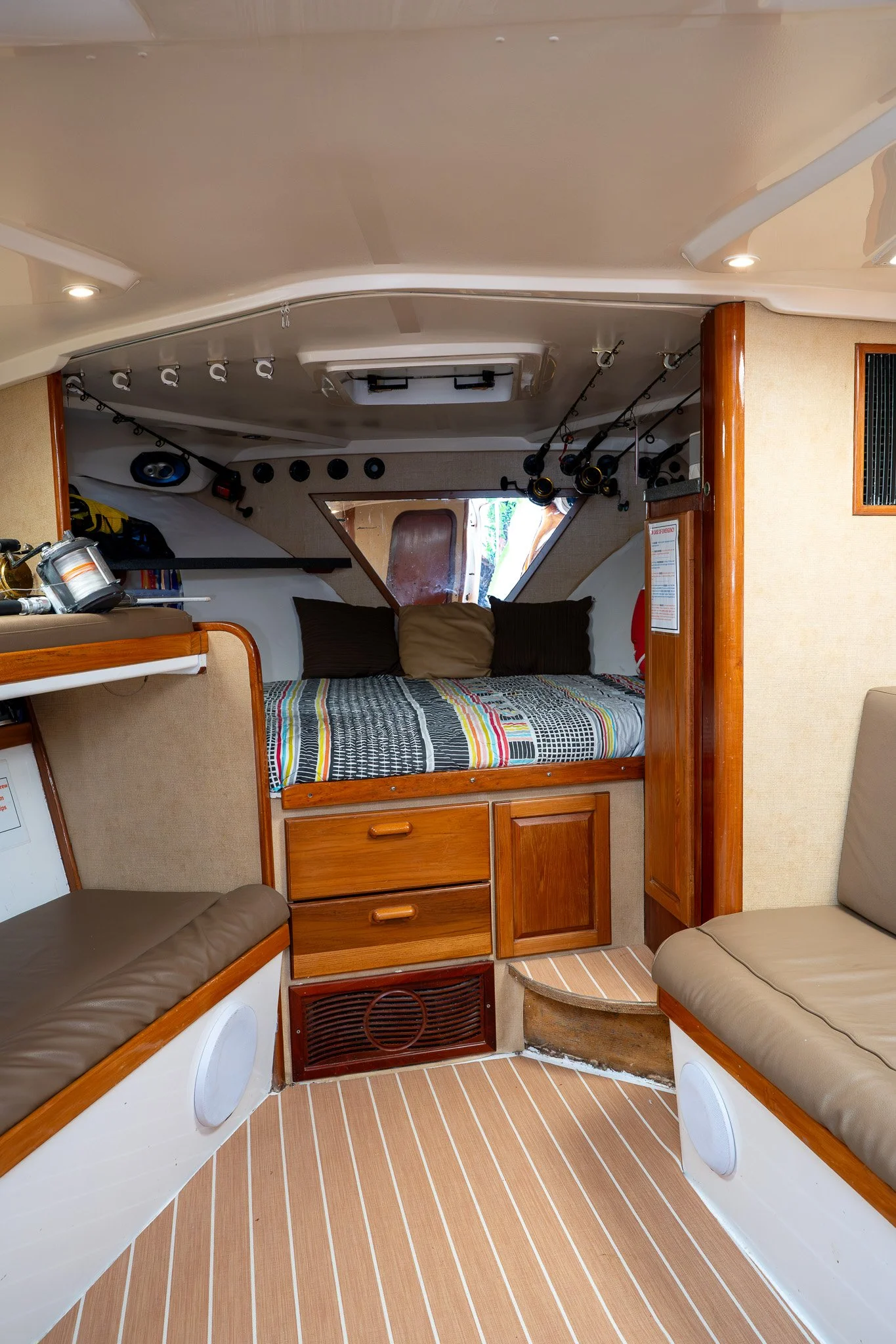 Interior of a boat cabin with a bed featuring a colorful striped bedspread, surrounded by wooden cabinetry and seating with beige cushions, and fishing rods mounted on the ceiling.