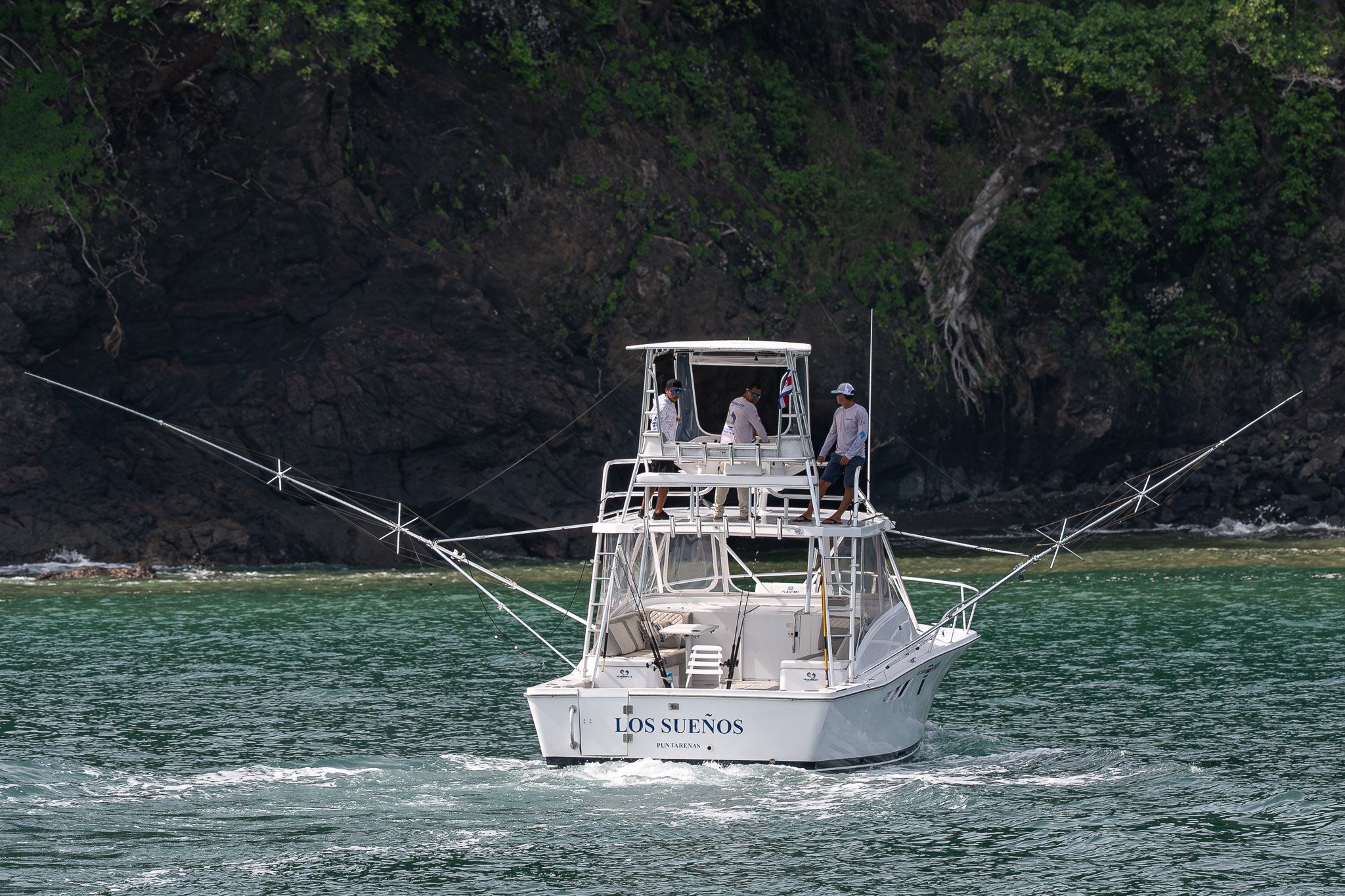 A white fishing boat named 'Los Sueños' on green water with a rocky cliff and trees in the background. Three people are on the upper deck of the boat, wearing casual clothing and hats.