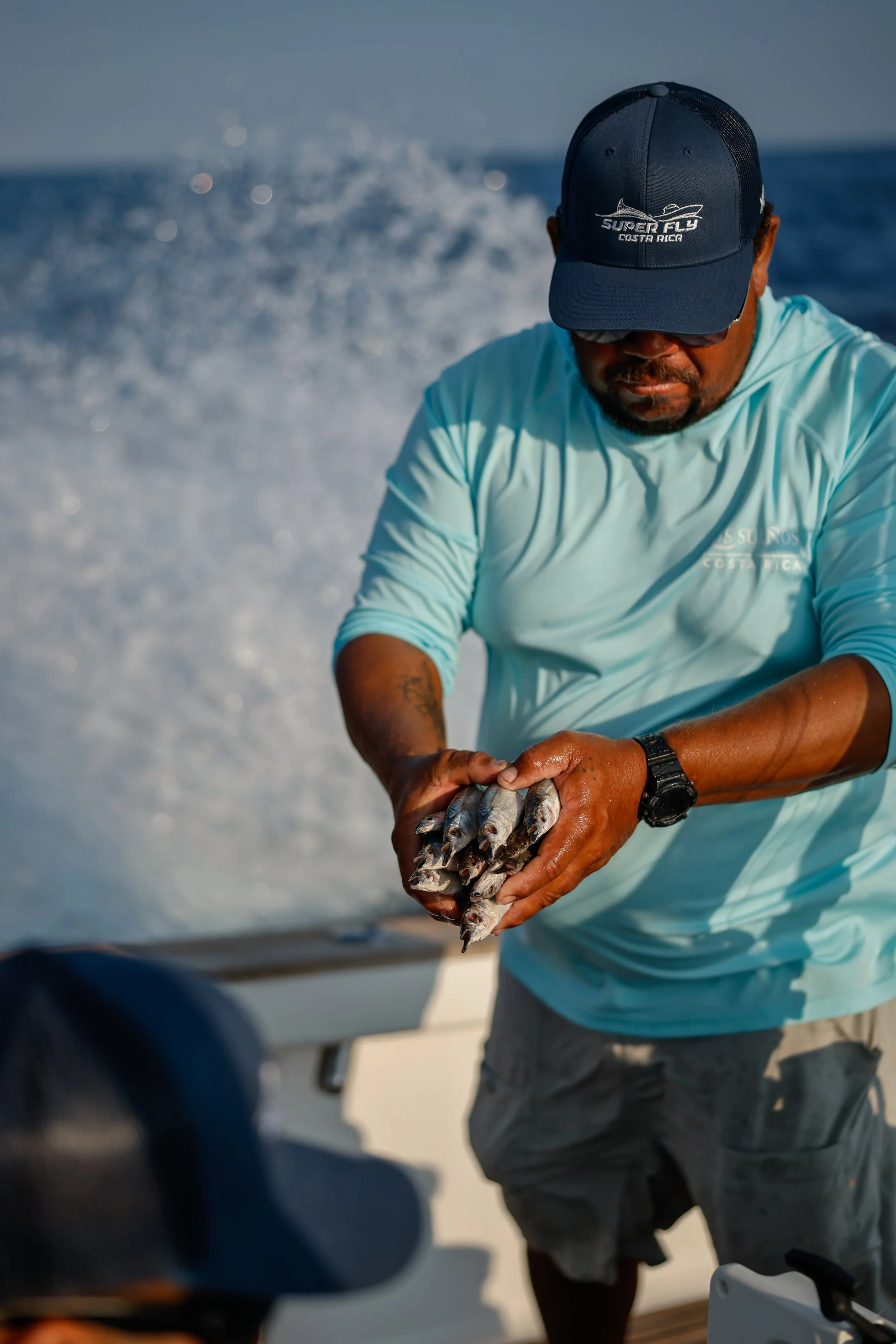 A man wearing a blue cap and light blue shirt holding small fish on a boat in the ocean.