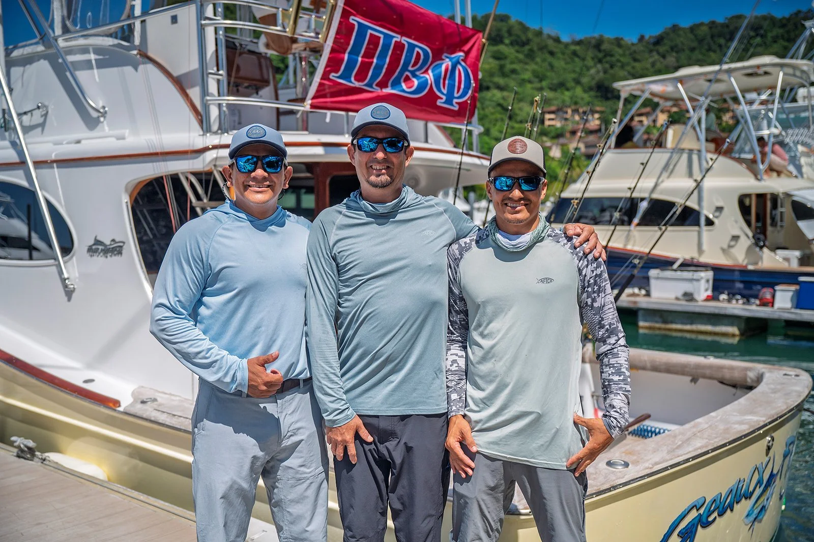 Three men standing on a dock in front of a boat, wearing casual outdoor clothing and sunglasses.