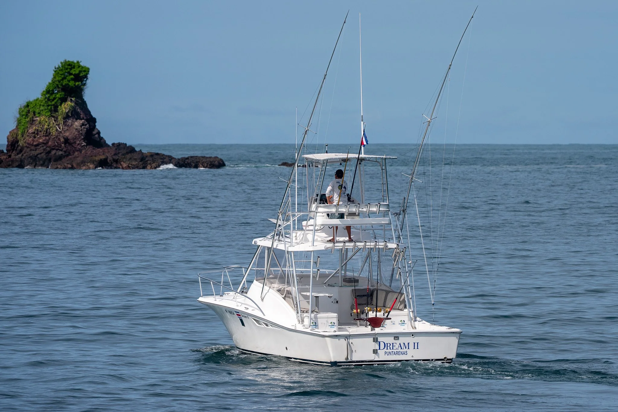 A white fishing boat named 'Dream II' sailing on the ocean, with a small island with greenery and rocks in the background, and a person onboard standing and observing.