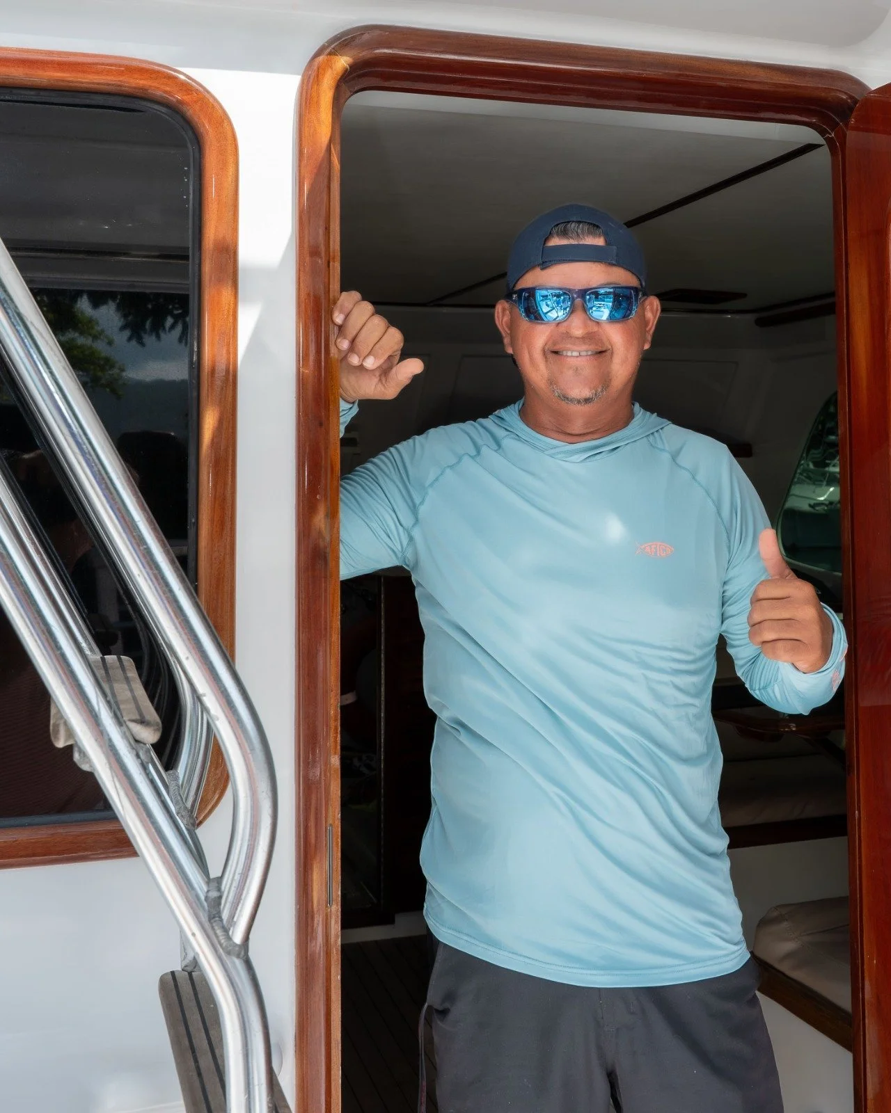 A smiling man wearing a blue long-sleeve shirt, sunglasses, and a backward cap stands at the entrance of a boat, giving a thumbs-up with his right hand.