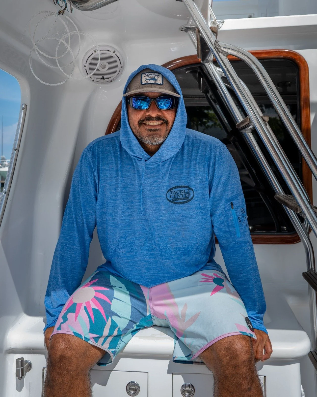 Man wearing sunglasses, a hoodie, and floral shorts sitting on a boat.