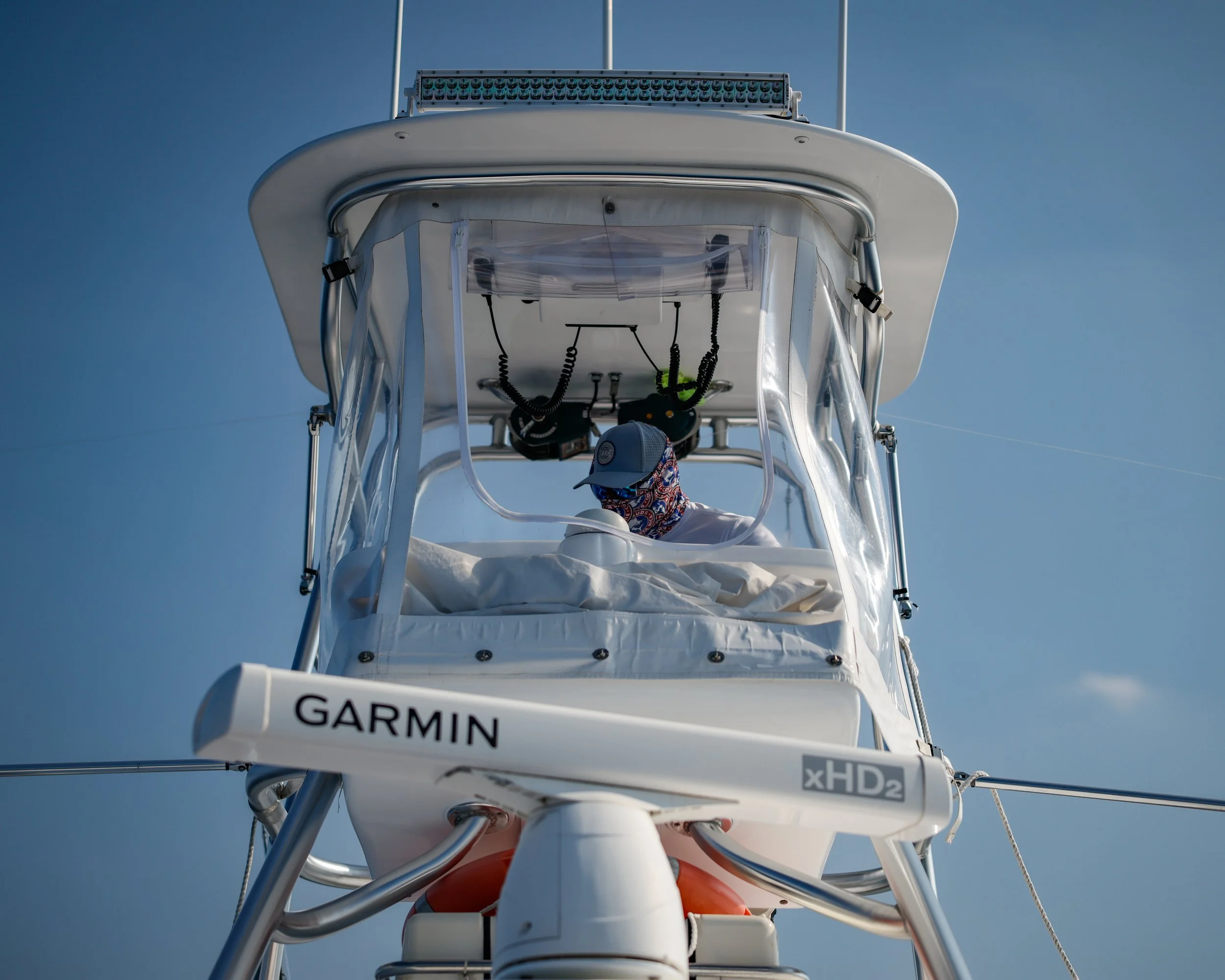 View from below of a boat with Garmin xHD2 equipment, a person wearing a cap and patterned face mask inside the cabin, against a background of a mostly clear sky with a few clouds.