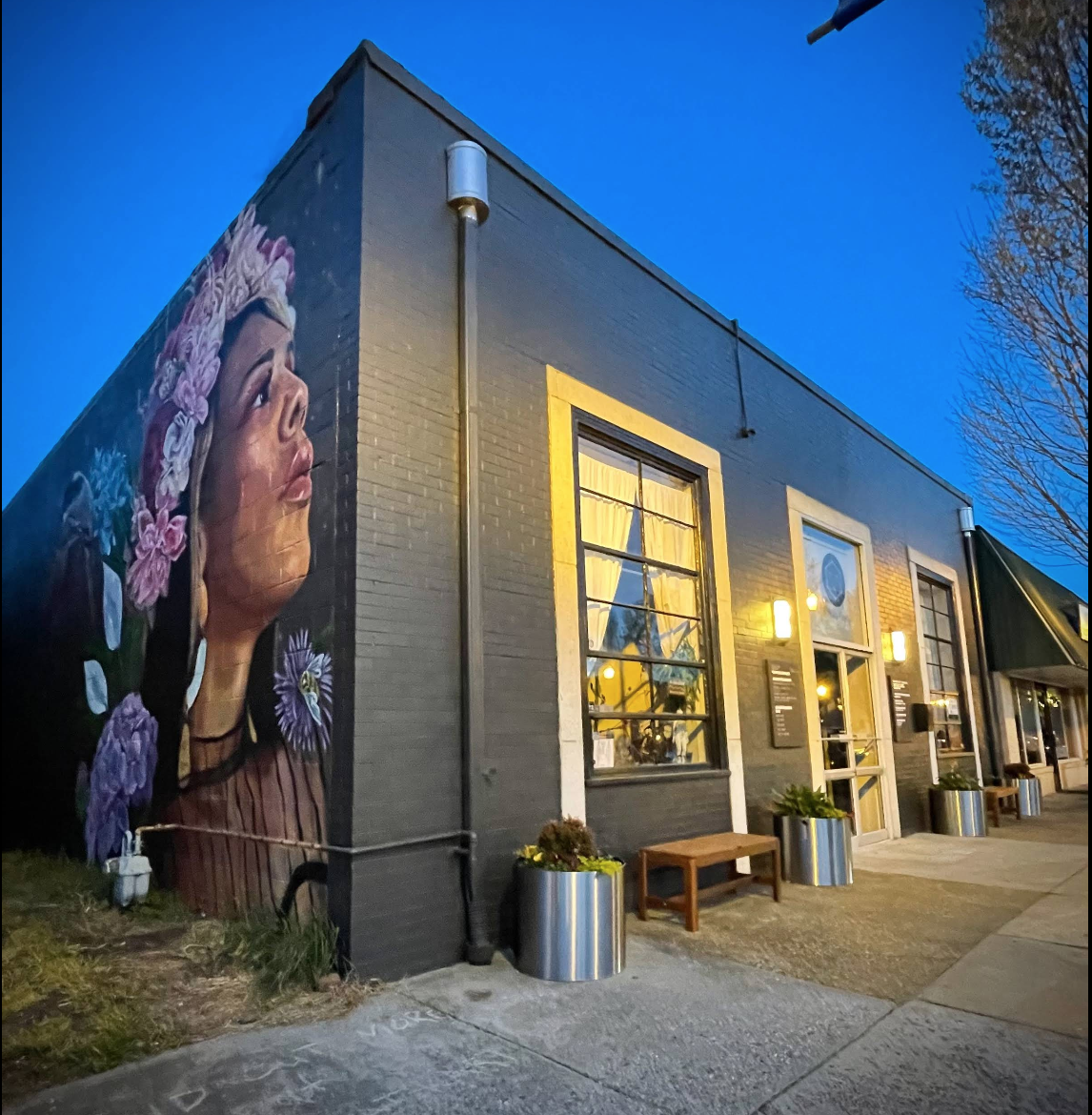 Private massage therapist office in Richmond VA. Street view of a building with a mural of a woman with flowers in her hair on the side wall, illuminated by exterior lights at dusk, with large windows and planters outside.
