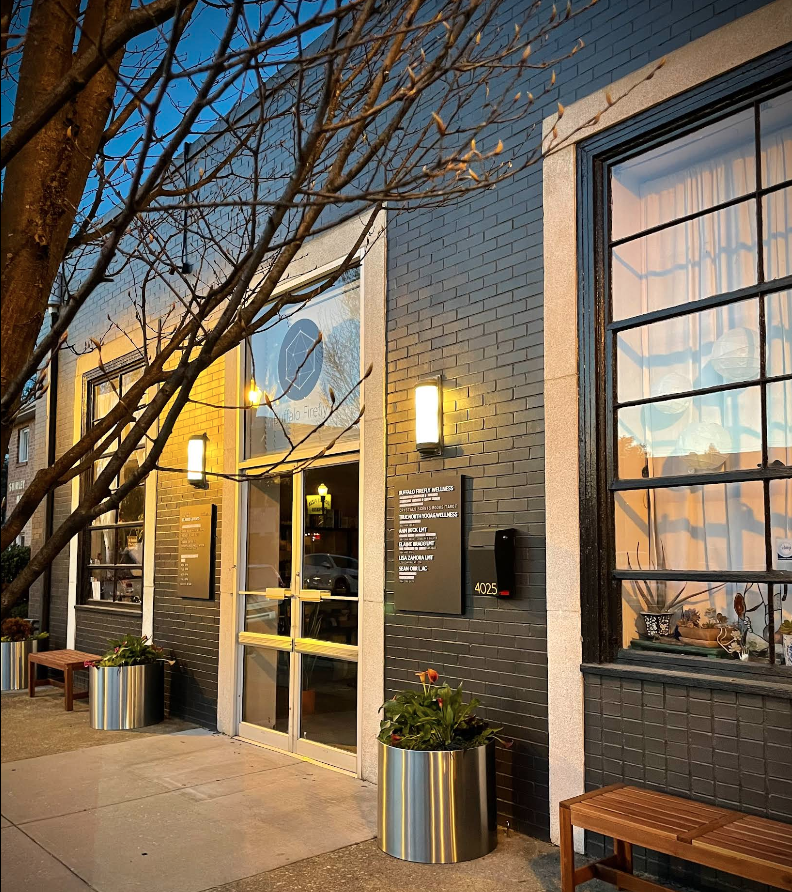 Massage therapist Richmond VA. Exterior of a modern building with a black brick facade, displaying windows and entrance door with a yellow sign, outside benches, and potted plants, during evening with warm lighting.