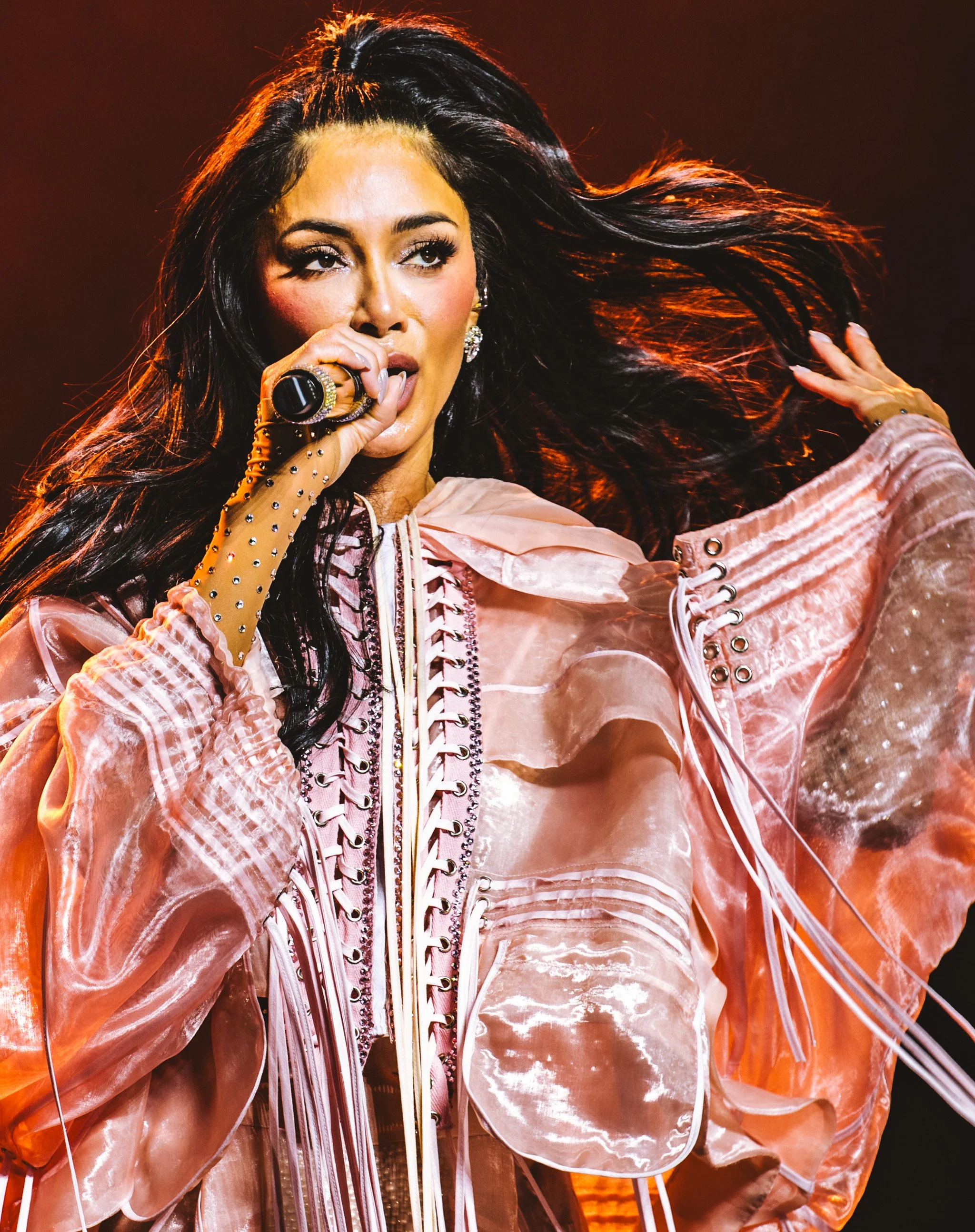A woman with long dark hair singing into a microphone, wearing a shiny pink jacket with lace-up details and chandelier earrings.
