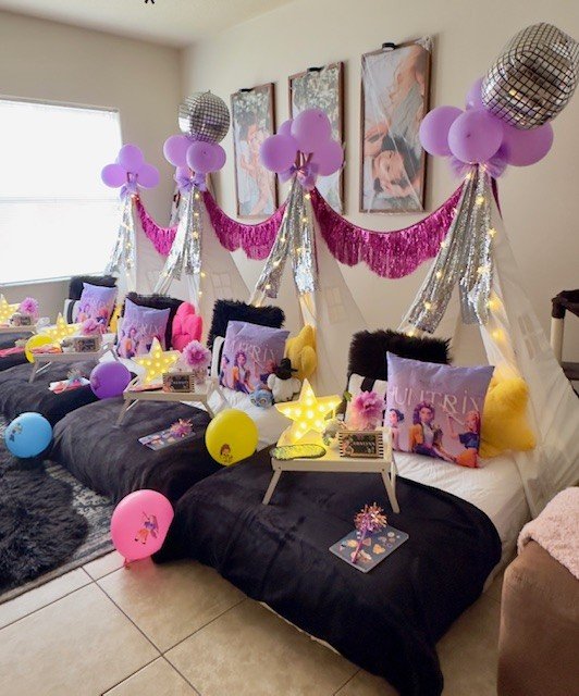 A living room decorated for a celebration with purple, pink, and yellow balloons, purple pillows with a character, and shiny silver and purple streamers.